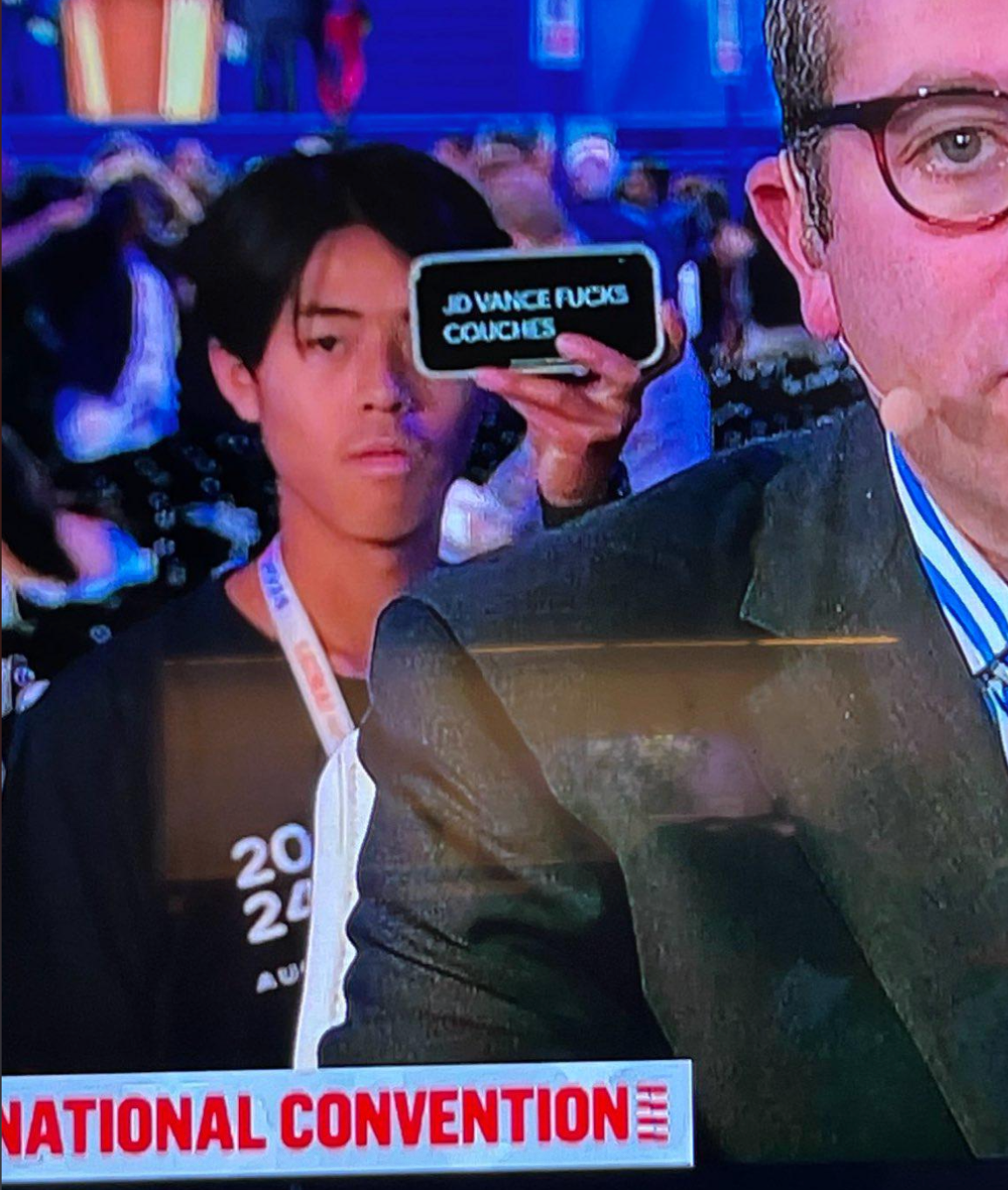 Person in a crowd holds up a phone with text, partially obscured. Event setting, possibly a convention