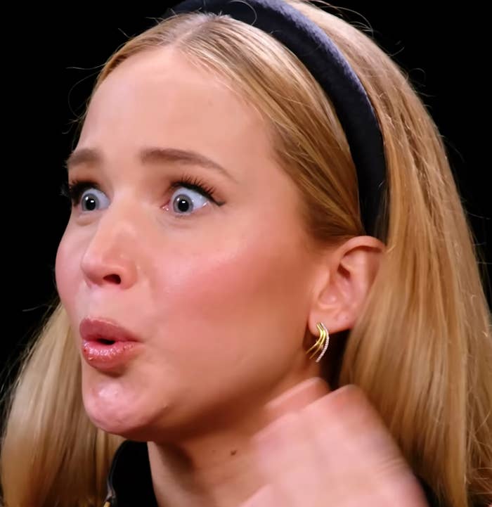 Person animatedly reacting with wide eyes and raised hands, wearing a headband and jacket, on a talk show