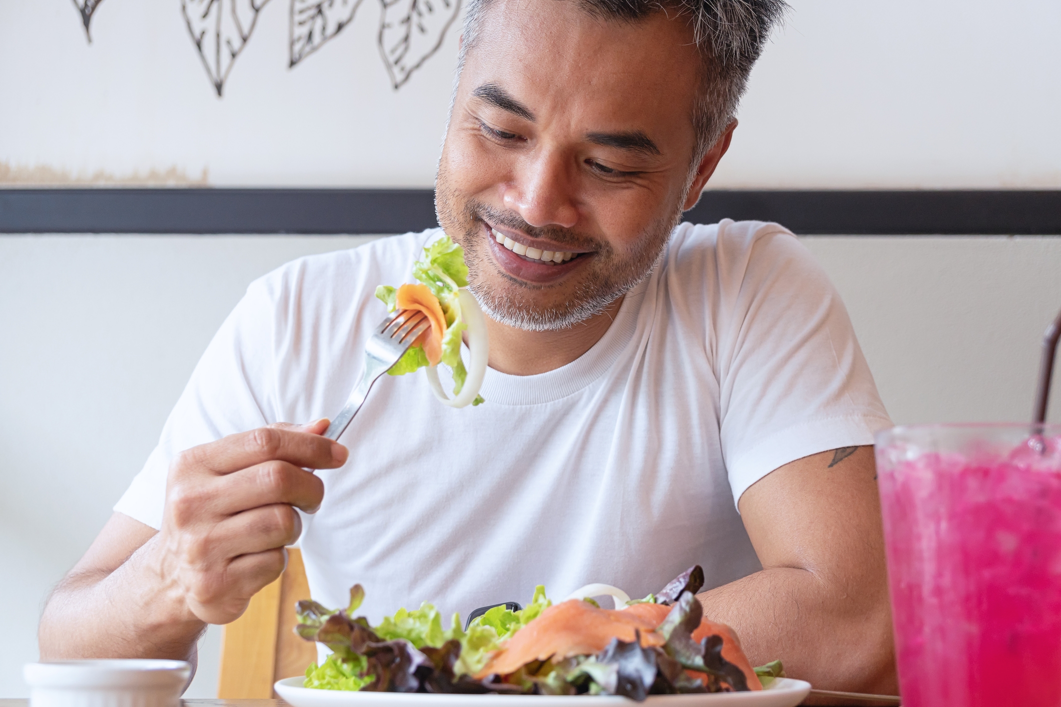 Person smiling while eating a salad at a table, holding a fork with lettuce and salmon