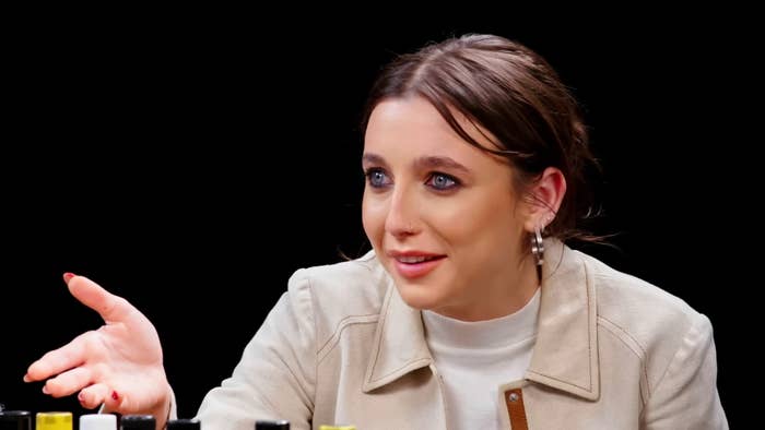 Person gesturing with open hand, wearing a light jacket and hoop earrings, sitting at a table with various sauces, likely in a show or interview setting