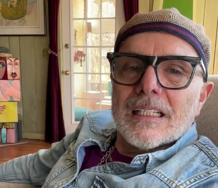 Persona con gafas y gorra sonríe, sentado en un sillón con cuadros artísticos y una puerta de fondo. Lleva camisa de mezclilla