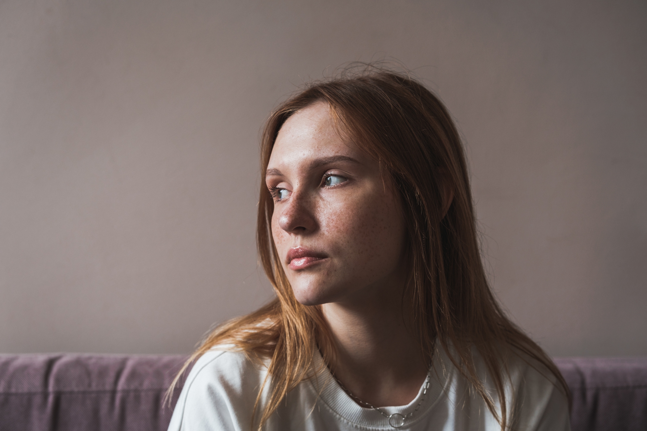 A person with long hair looks to the side, seated on a couch. Neutral expression and casual attire