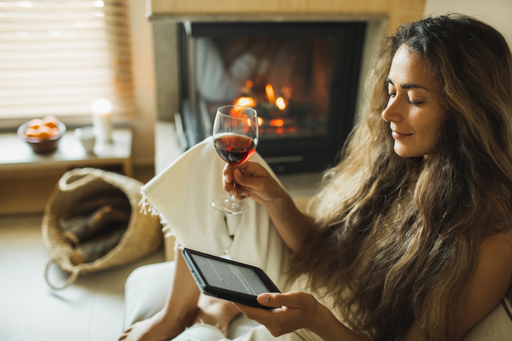 Person relaxing by a fireplace, reading on an e-reader and holding a glass of wine, with a basket of logs nearby