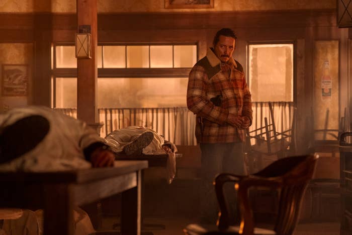 Hombre en ambiente rústico con camisa a cuadros observa cuerpos en una mesa, escena de suspenso o crimen