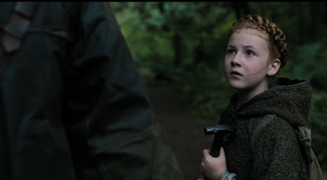 Niña con trenza y sudadera con capucha sostiene herramienta en un bosque, mirando hacia un adulto