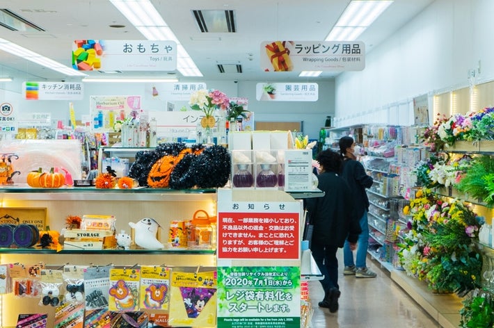 ダイソー店内、ハロウィーングッズと花が並ぶ。顧客が買い物中。