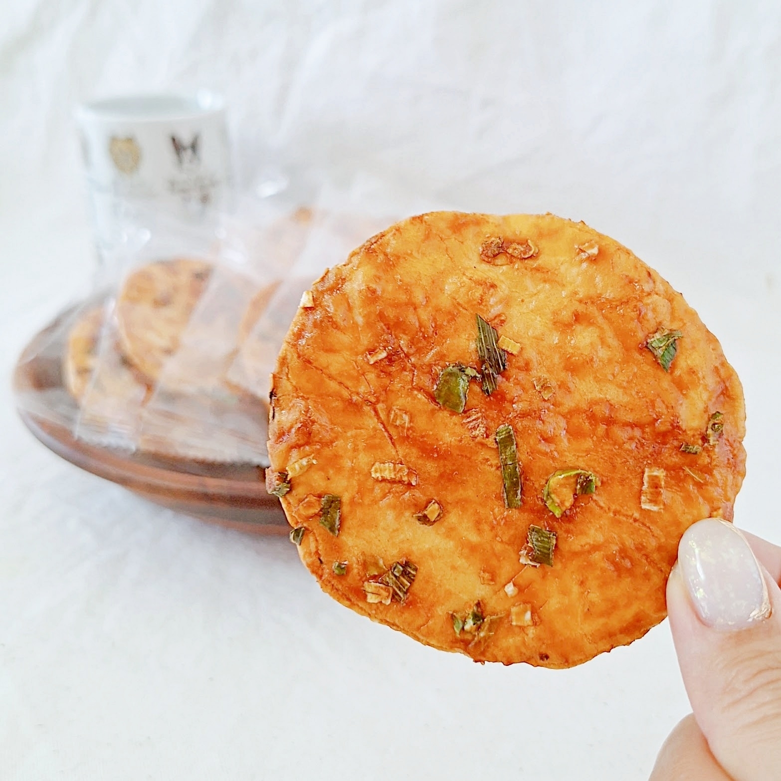 手に持たれたせんべい。背後には木製の皿に盛られた他のせんべいとカップがあります。