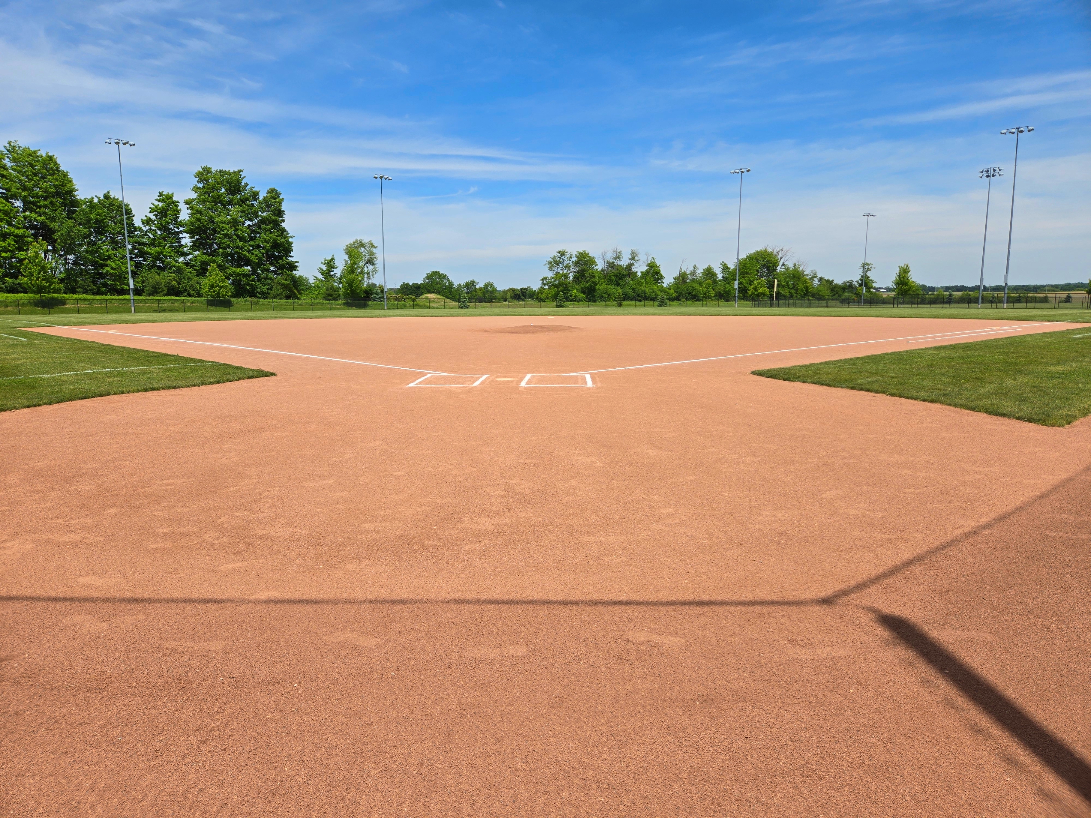 Empty baseball field with bases and pitcher's mound under a clear sky, surrounded by lush trees in the distance