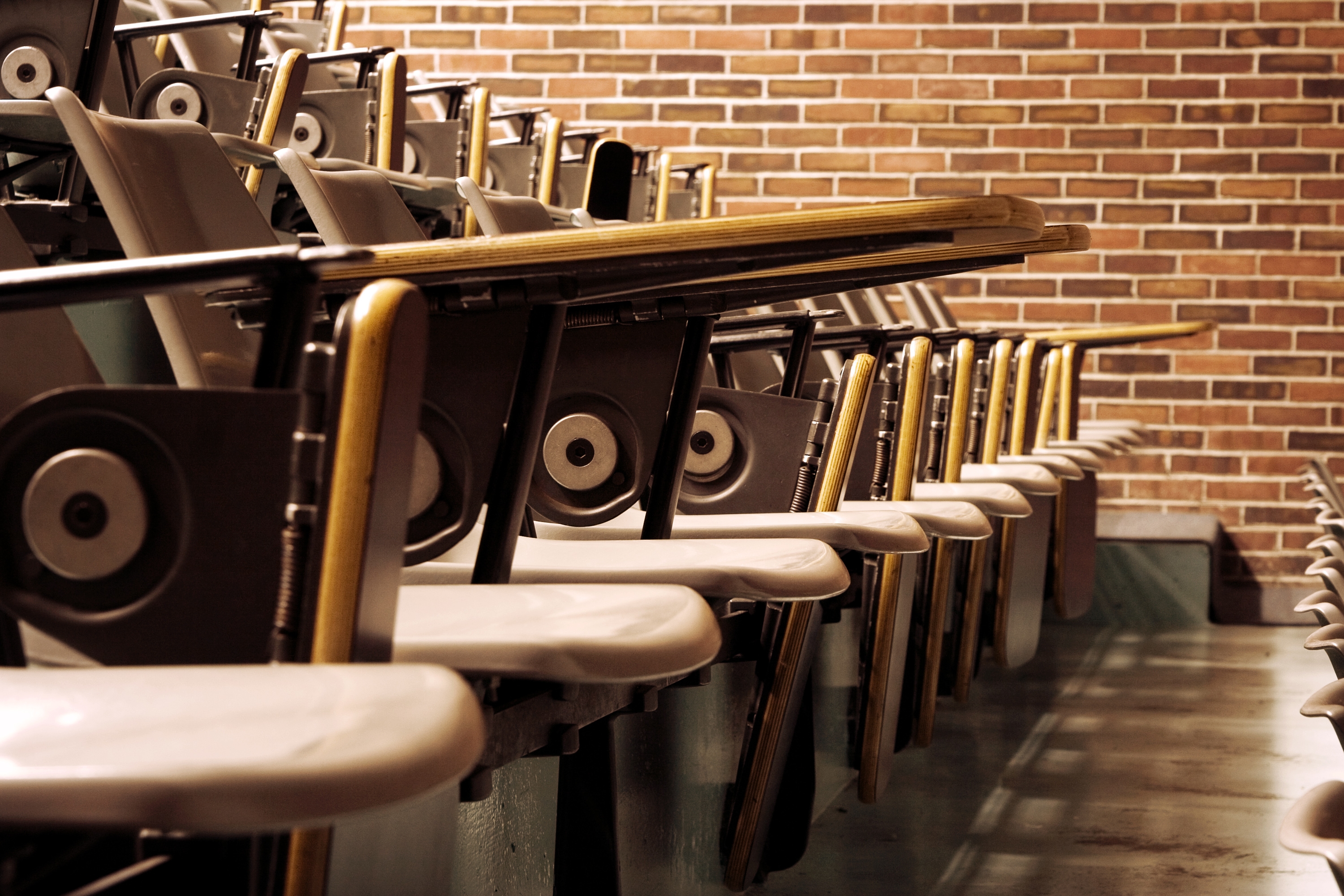 Empty lecture hall with rows of wooden seats and foldable desks, set against a brick wall backdrop