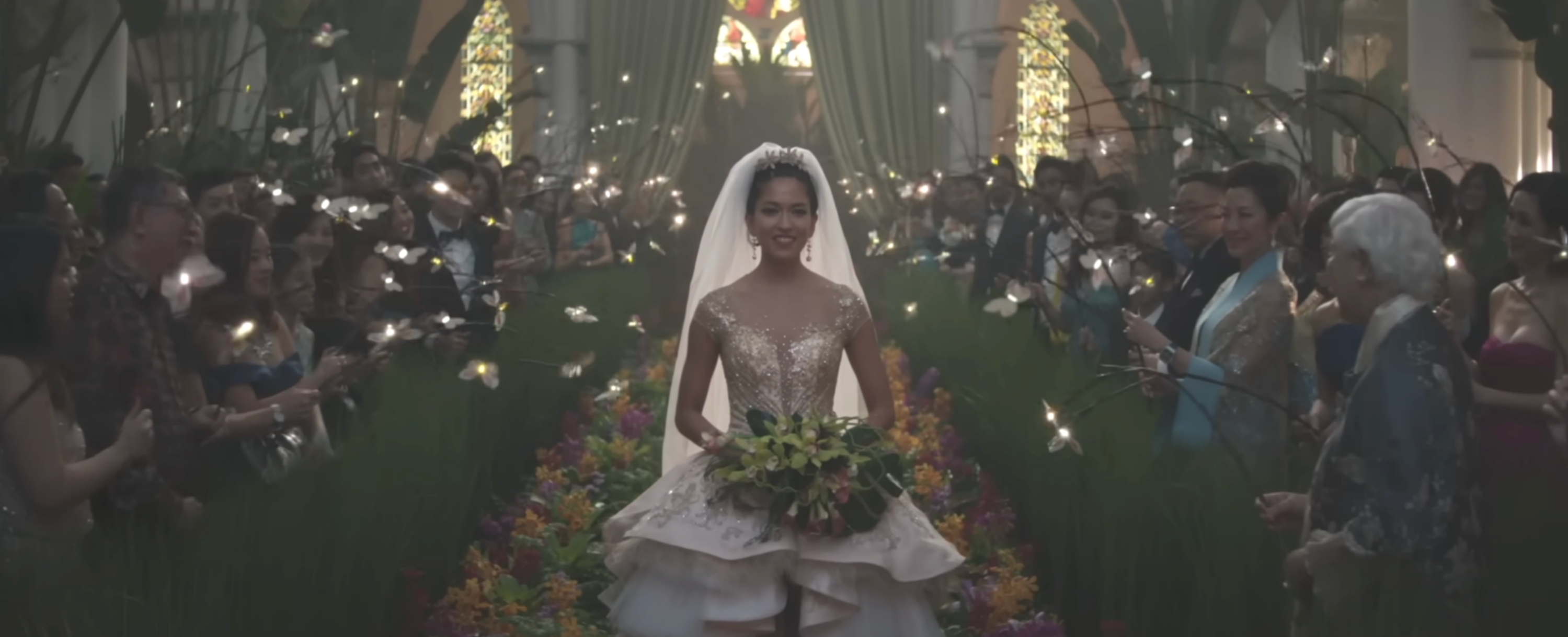 Bride in a gown with floral accents walks down an aisle adorned with lights and greenery, surrounded by guests
