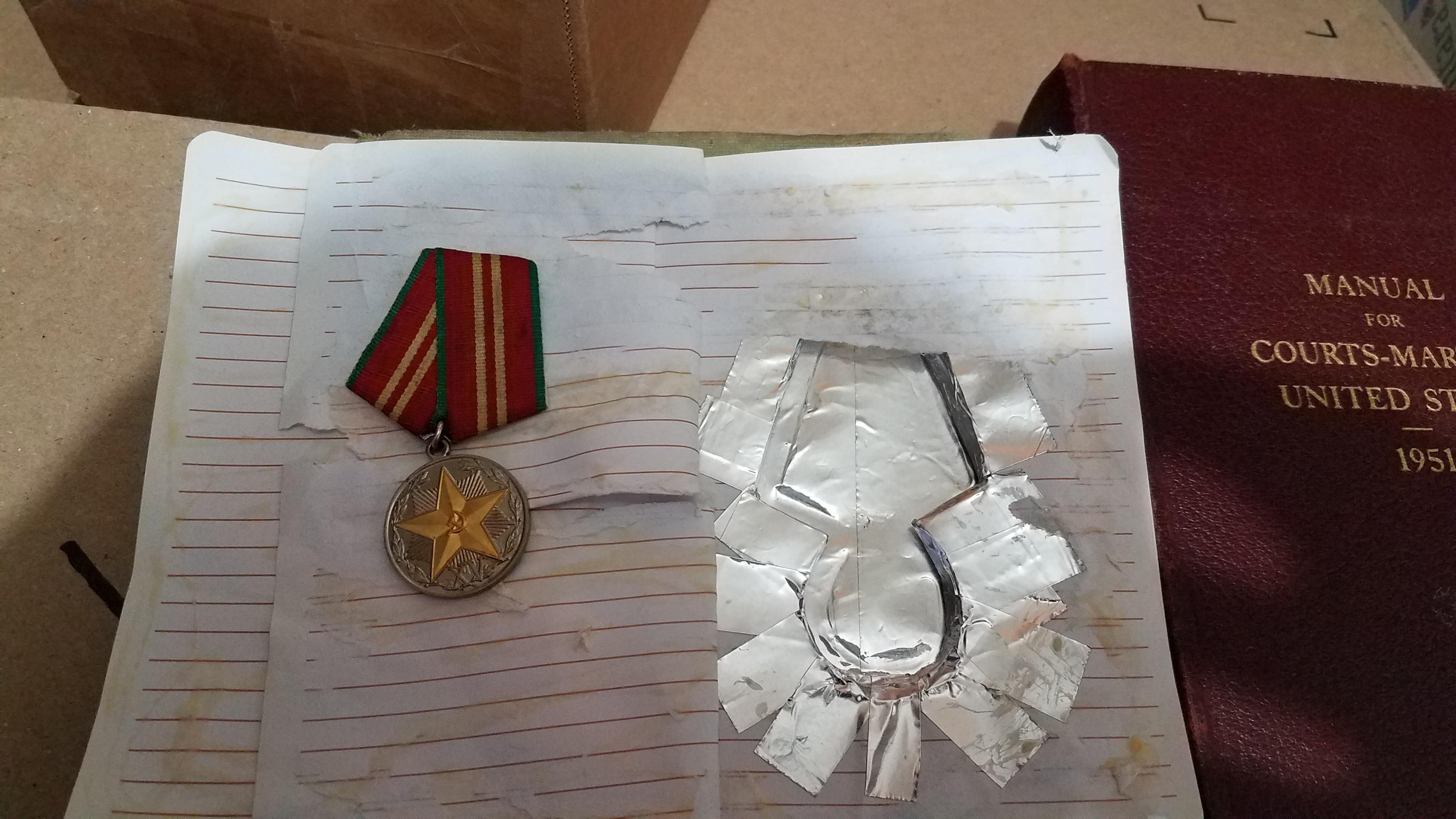 A military medal with a star and ribbon is placed on handwritten notes next to a damaged foil pouch inside a book. A red book titled "Manual for Courts-Martial 1951" is nearby