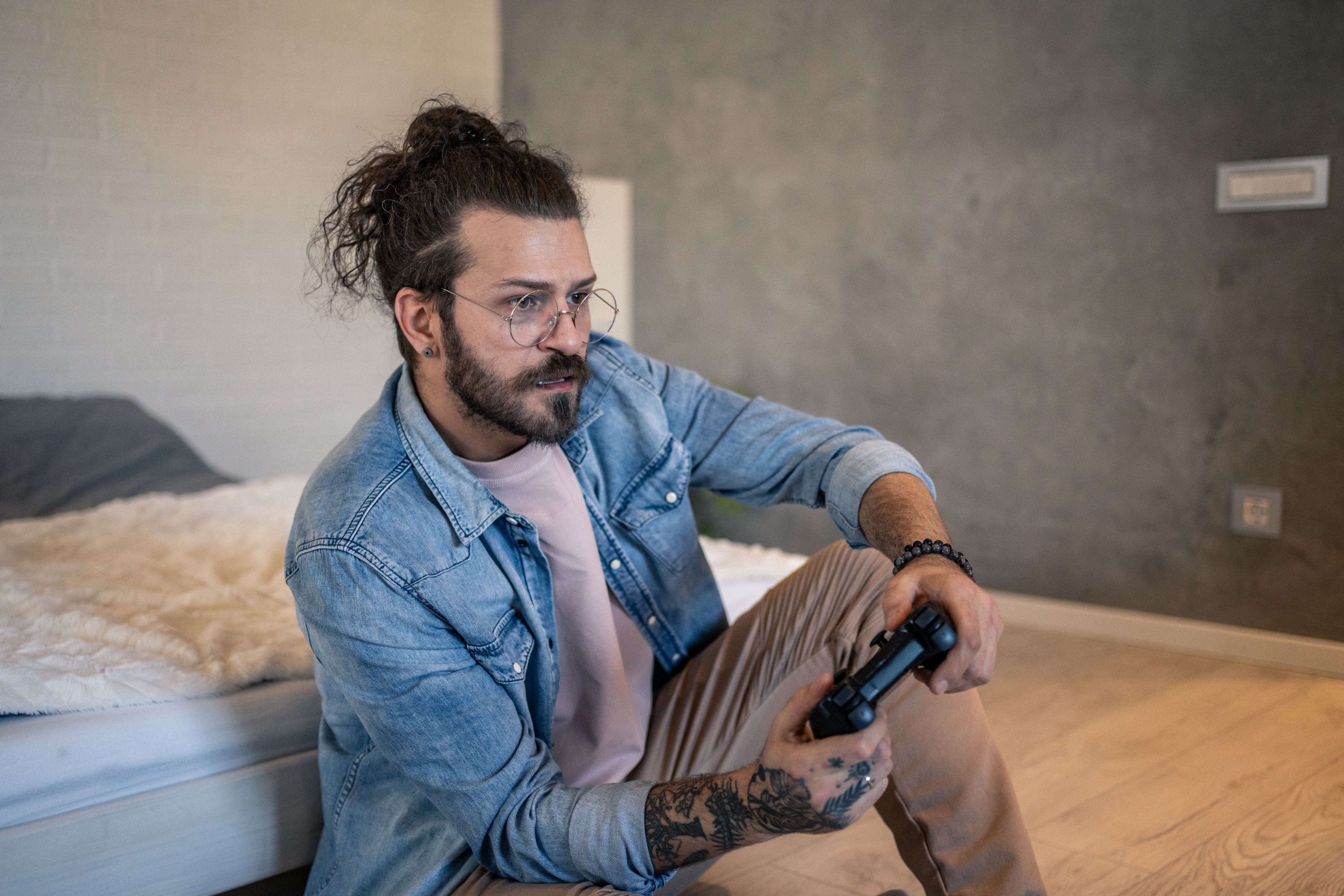 Person with glasses and tattoos playing video games in a casual indoor setting, seated on a bed