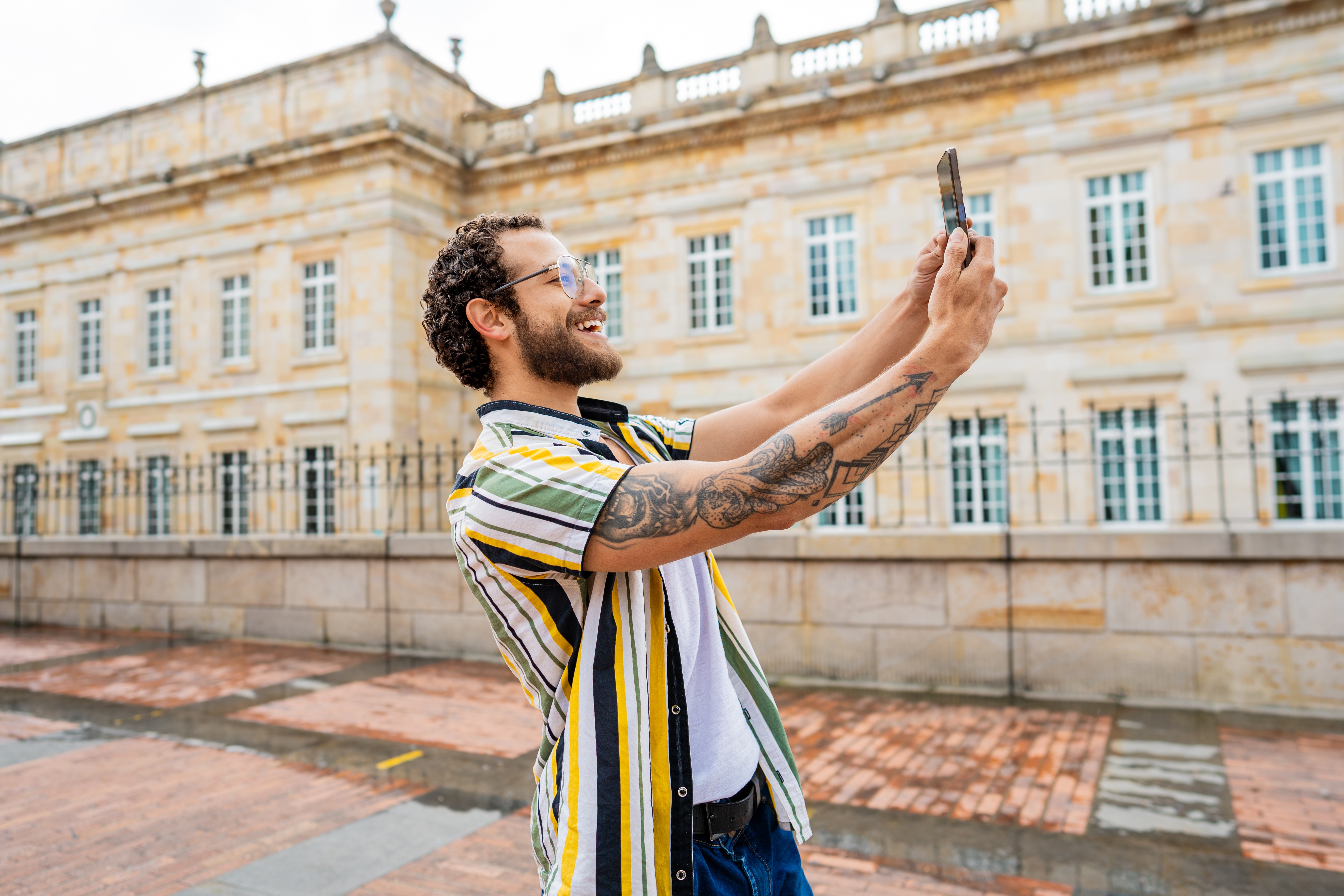 Person taking a selfie outdoors in front of an elegant building, smiling, wearing a striped shirt and glasses