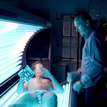 Person in a tanning bed holding a magazine, while an older man stands beside them holding a coffee mug, inside a dimly lit room