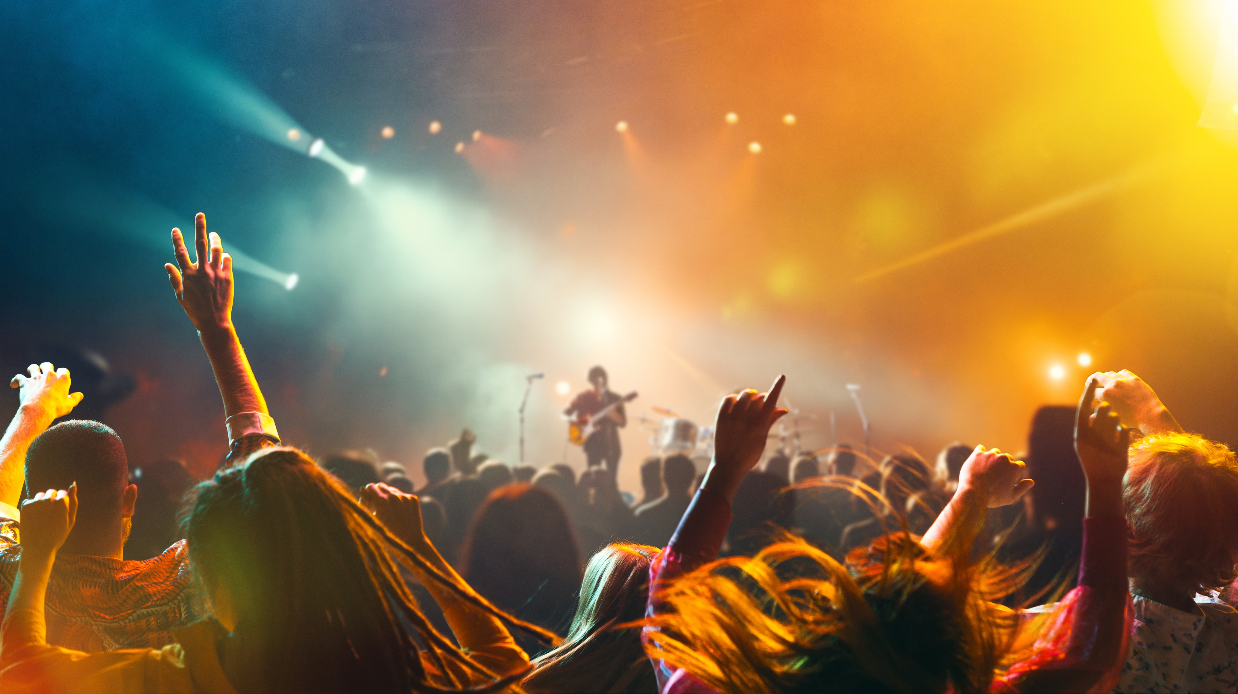 A crowd enjoys a live music concert, with a guitarist performing on stage under bright lights. The audience is energetically dancing and cheering