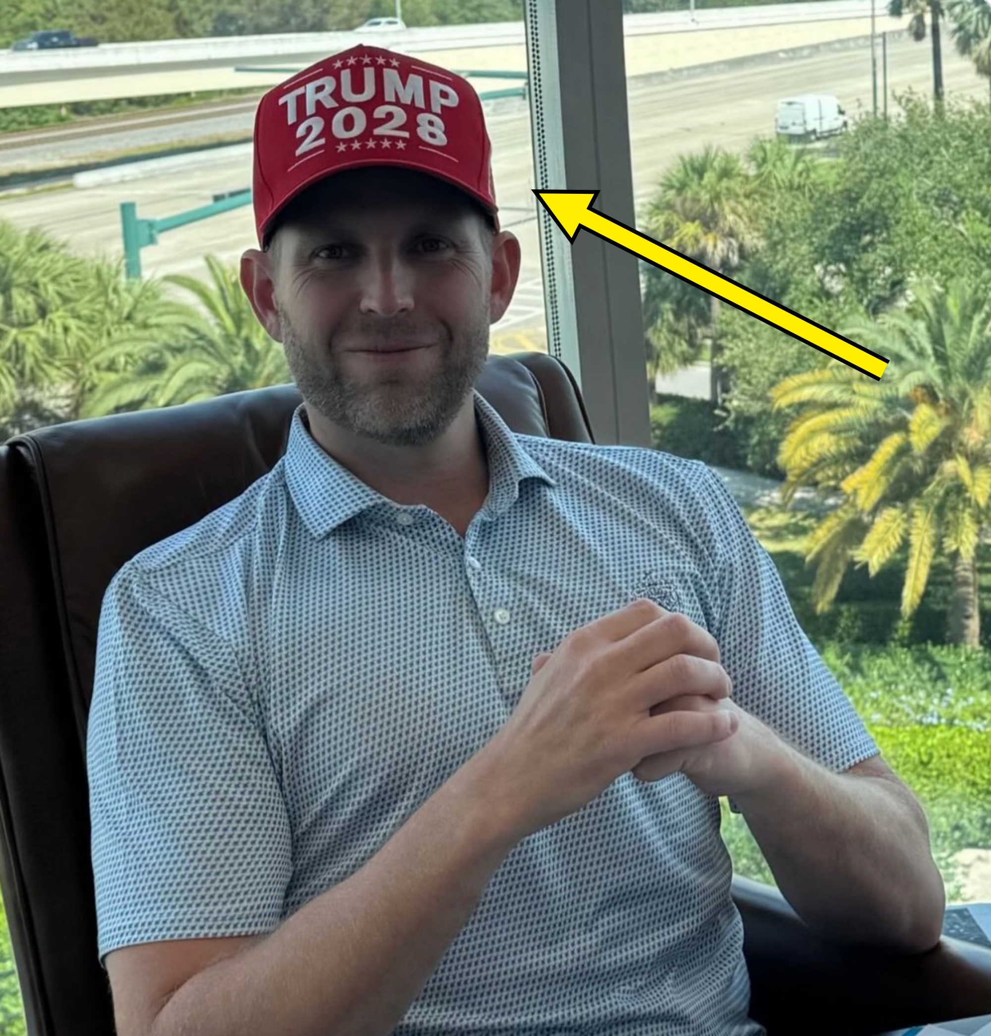 A person sits in a chair by a window, wearing a "Trump 2028" hat and a patterned polo shirt, with palm trees visible outside