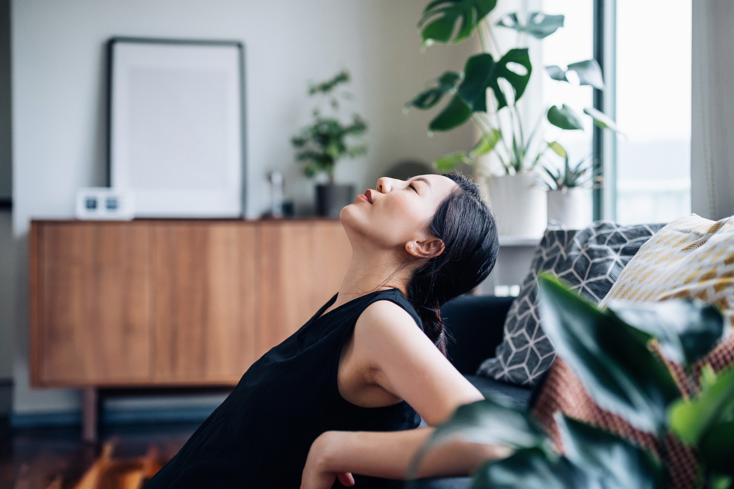 Person relaxing on a couch, leaning back with eyes closed, surrounded by indoor plants and modern decor. Peaceful home setting