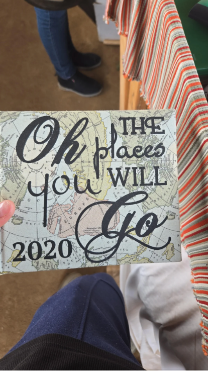 Hand holding a book that says "Oh, the places you will go 2020" on the cover overlaid on a world map
