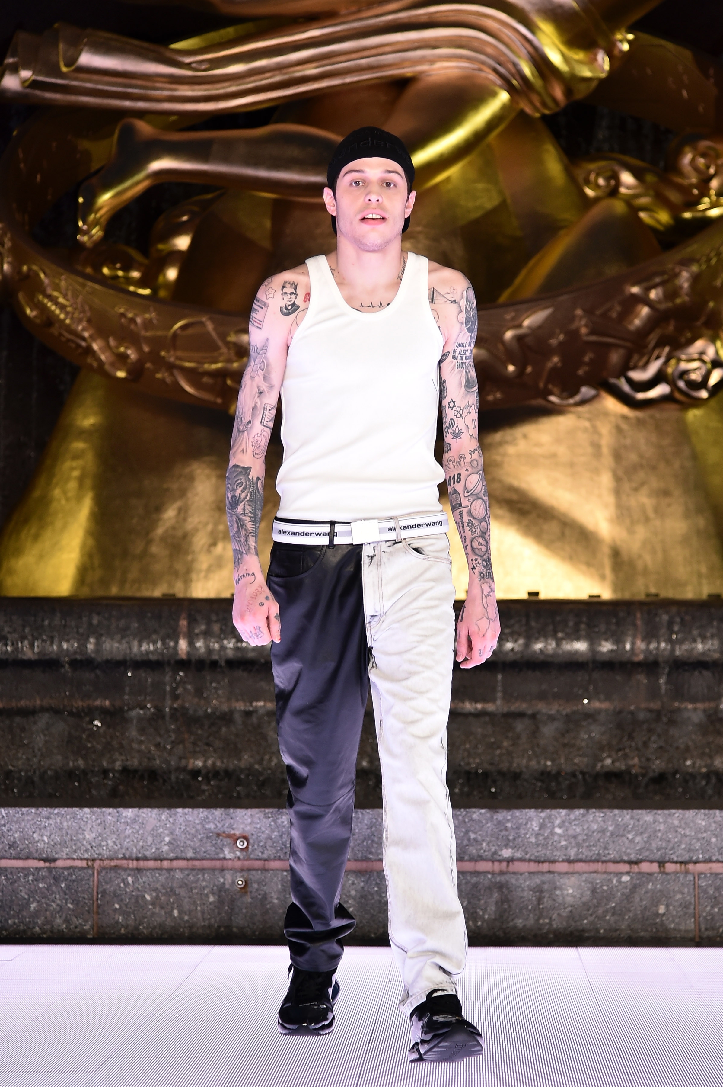 Person with tattoos, wearing a white tank top and two-tone pants, walks on a runway with a golden sculpture in the background