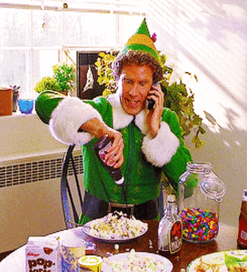 Person in an elf costume enthusiastically adds syrup to a spaghetti dish while talking on the phone. Playful holiday scene with candy and soda nearby