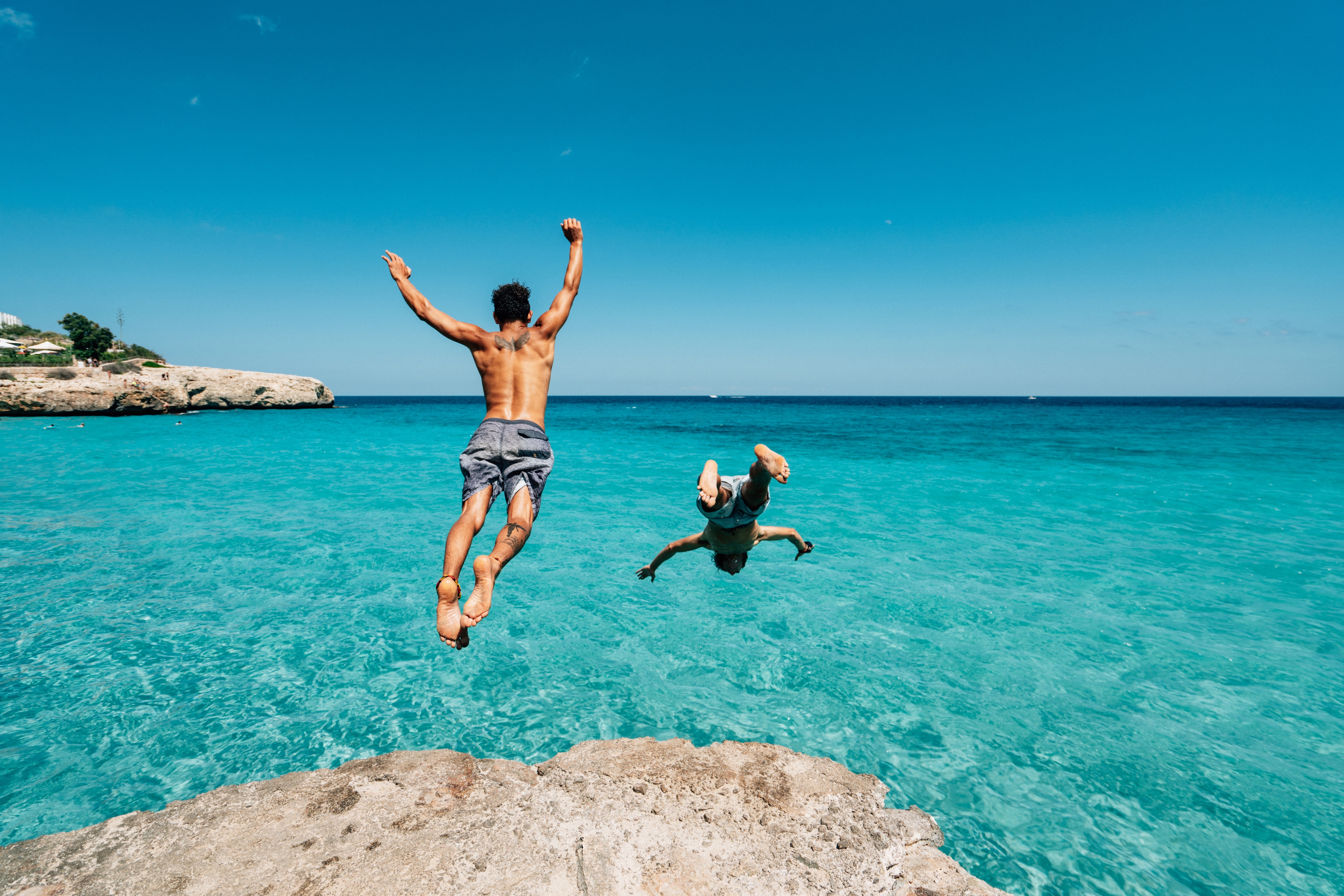 Two people jumping into the ocean from a rock, with arms raised joyfully, under a clear sky