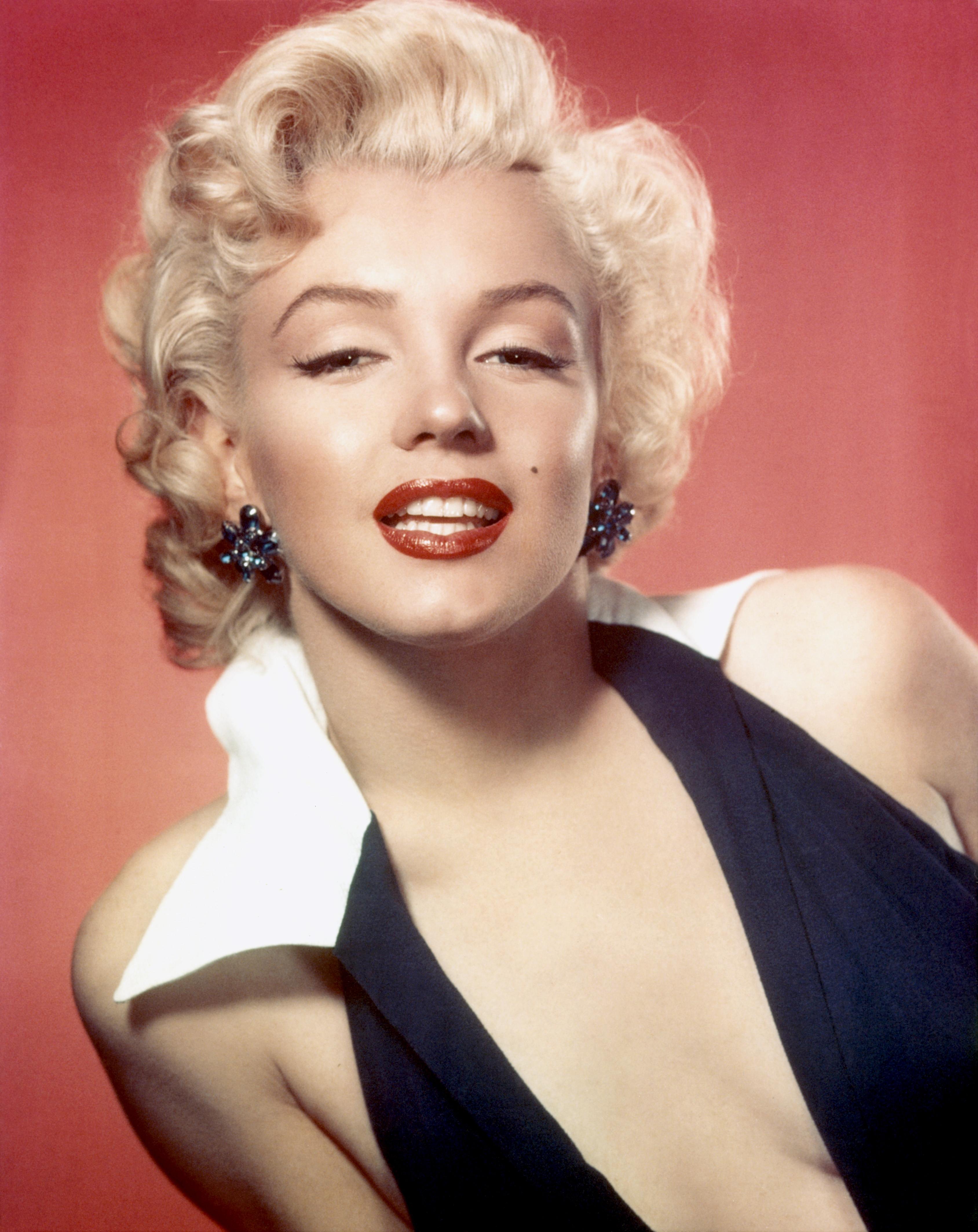 Person posing with classic Hollywood glam style, featuring curled hair, bold lipstick, and elegant halter-neck dress with statement earrings