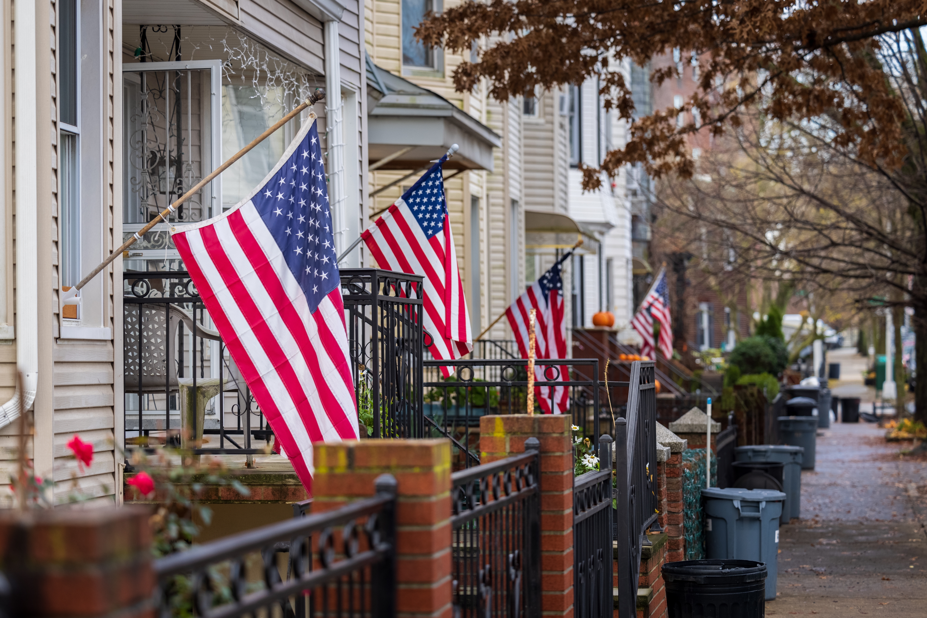 American flags displayed on multiple residential porches along a quiet neighborhood street lined with trees and brick fences