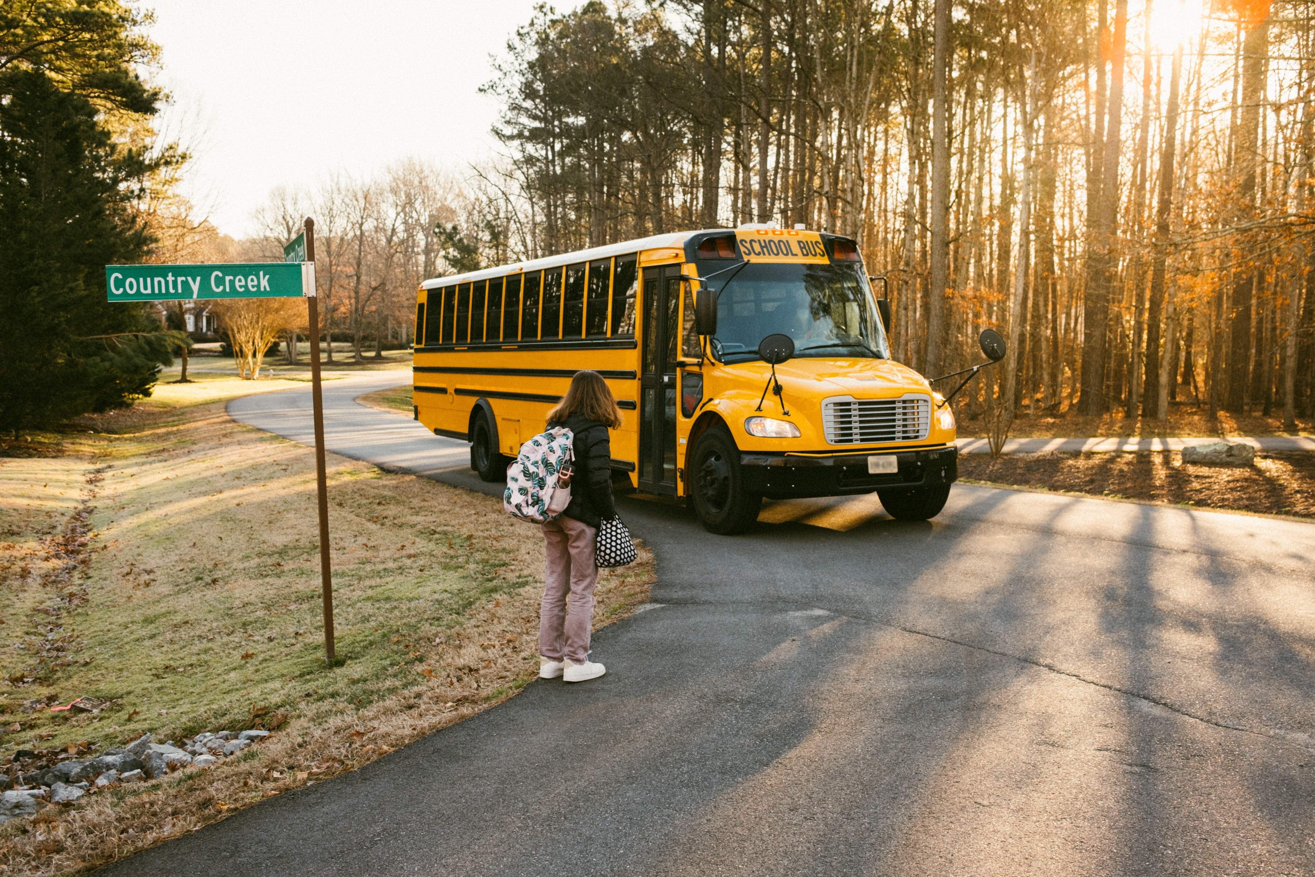 A student with a backpack waits for a school bus at a rural crossroads marked "Country Creek" during sunrise