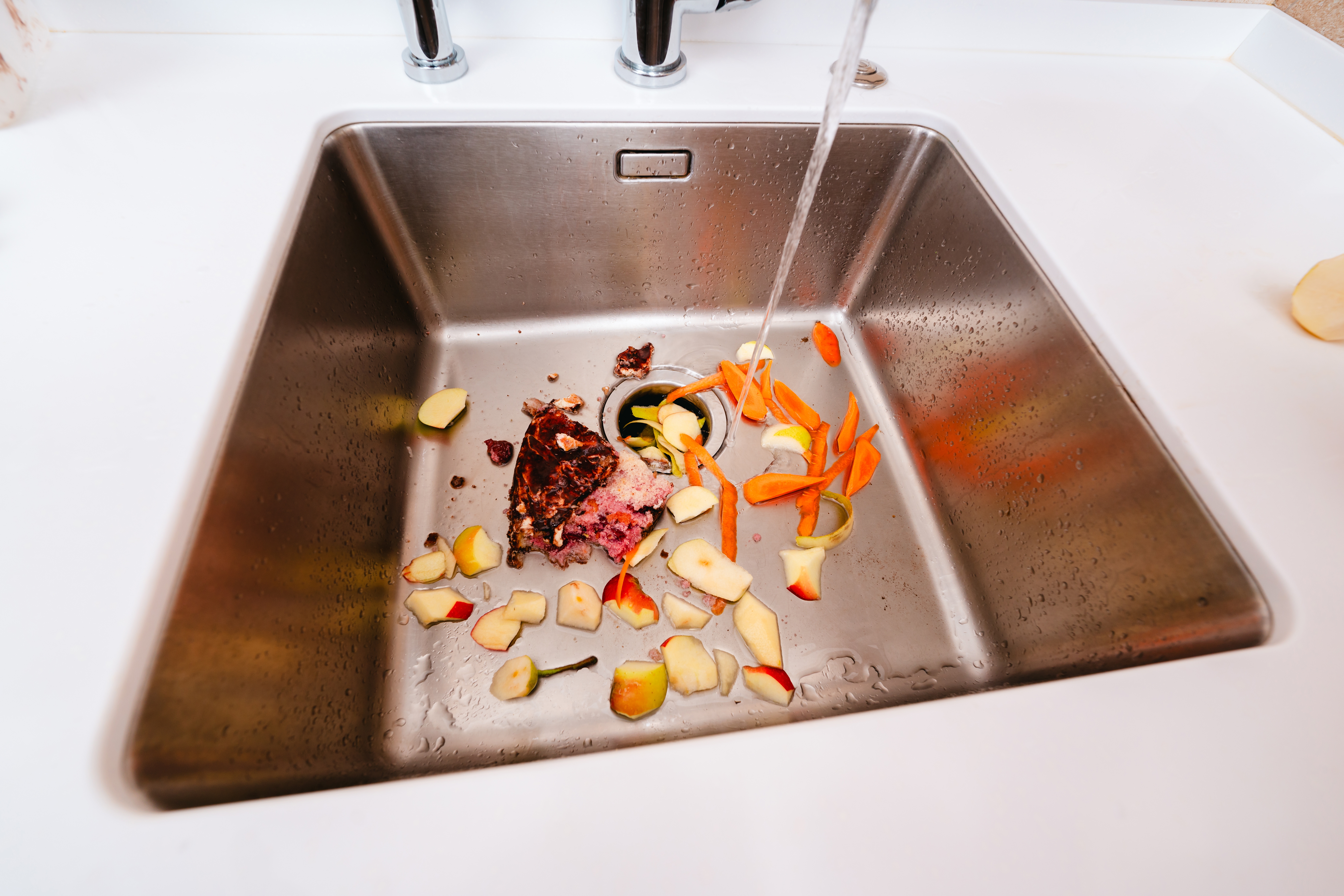 Food scraps including carrots and apple peels in a kitchen sink with running water
