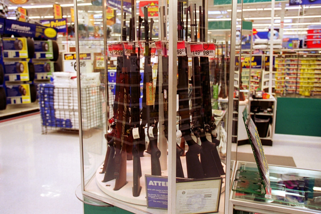 Glass display case with rifles in a store. Price tags are visible. Shelves of merchandise are in the background