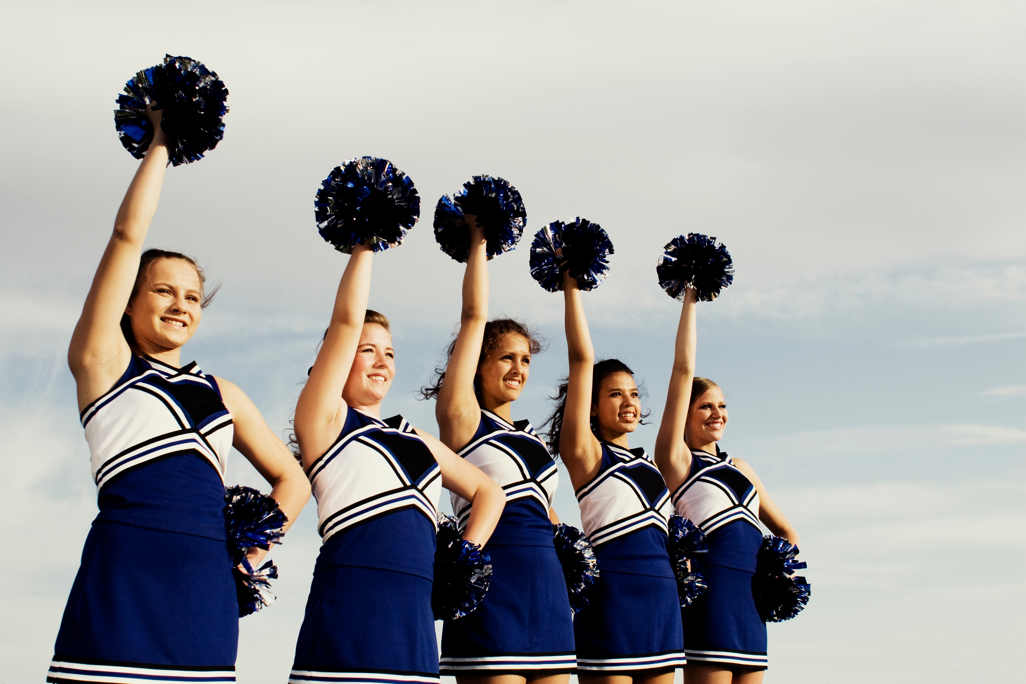 Five cheerleaders perform outdoors, holding pom-poms aloft in coordinated blue and white uniforms