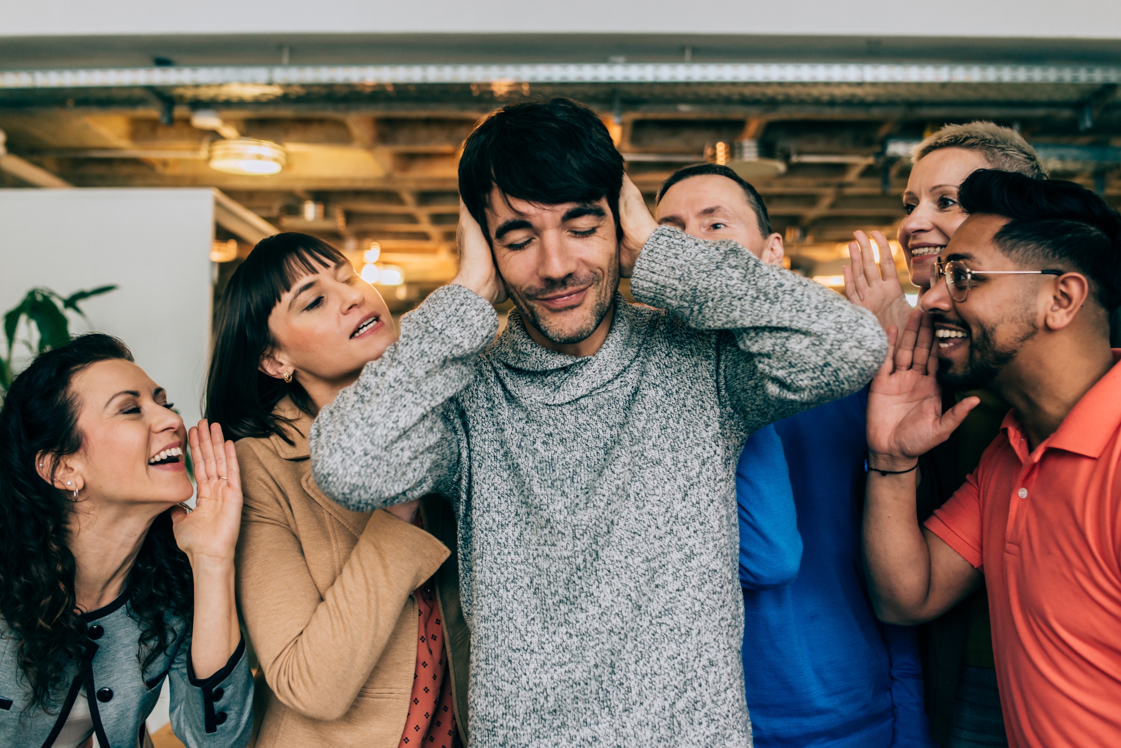 A person covering their ears stands amid a group of people surrounding them, appearing to whisper or shout