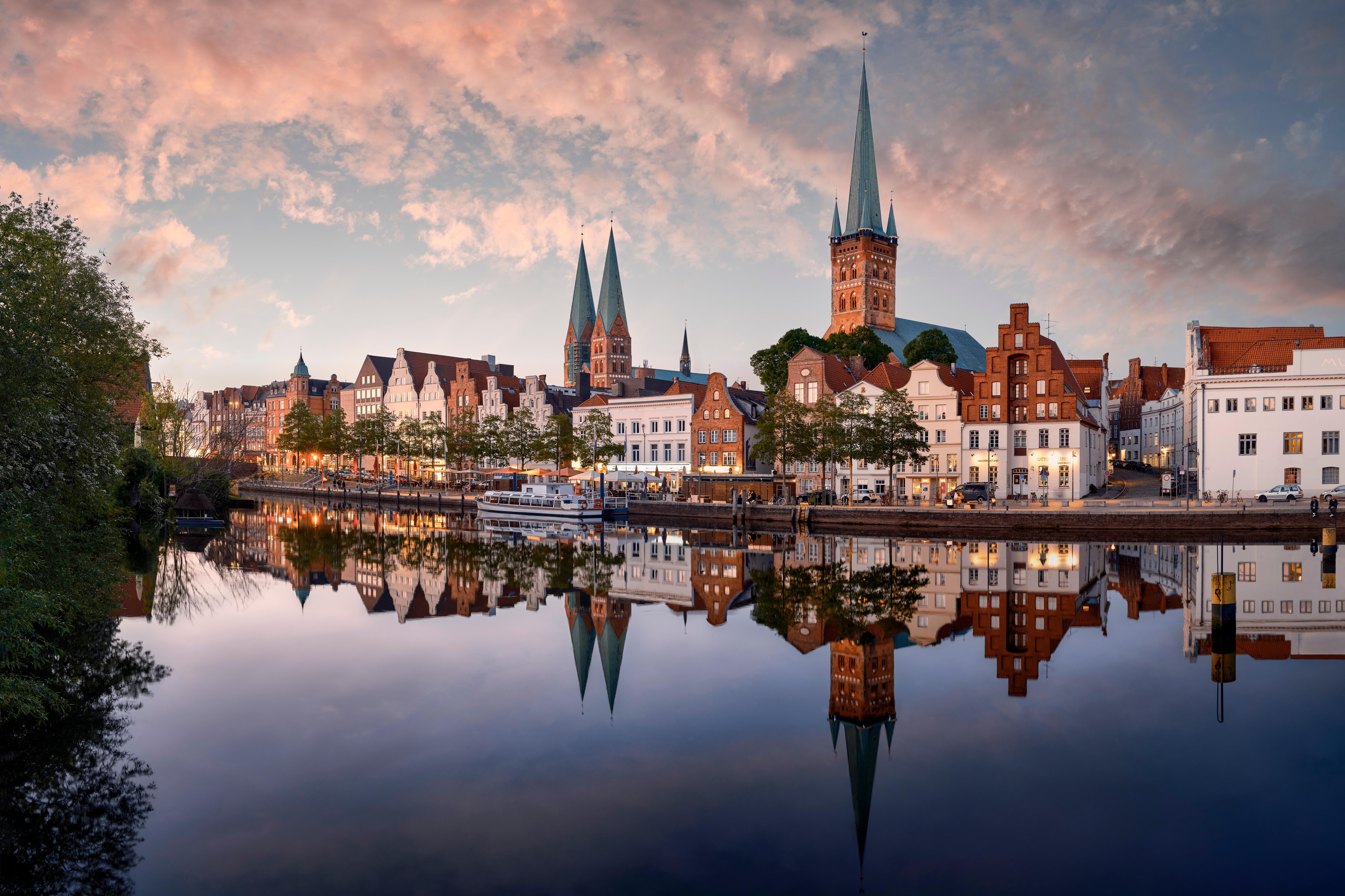 Historic Lübeck skyline reflecting in calm river, featuring medieval architecture with prominent church spires under a partly cloudy sky