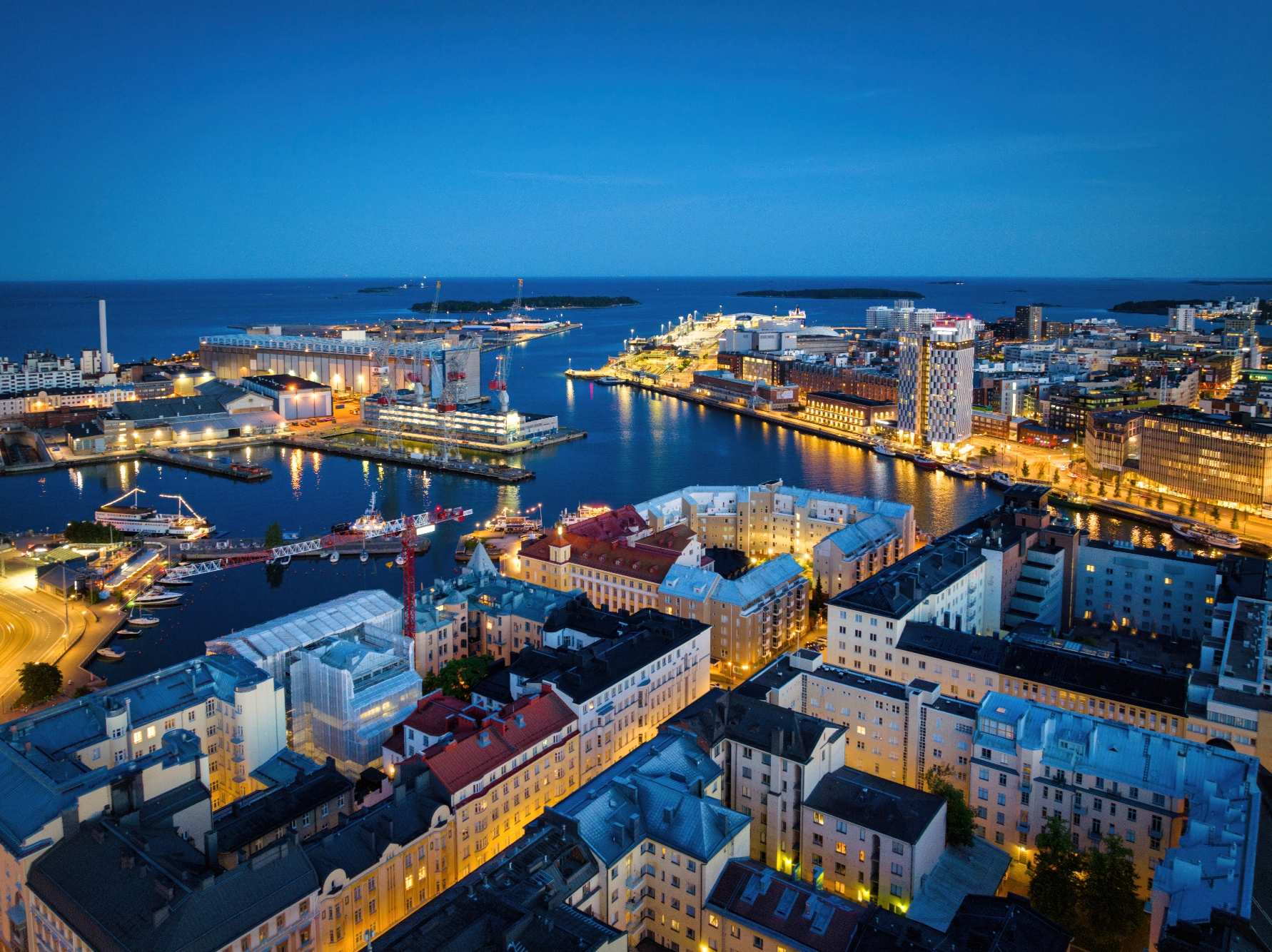 Aerial view of a city waterfront at night, showcasing illuminated buildings and boats, with a clear evening sky in the background