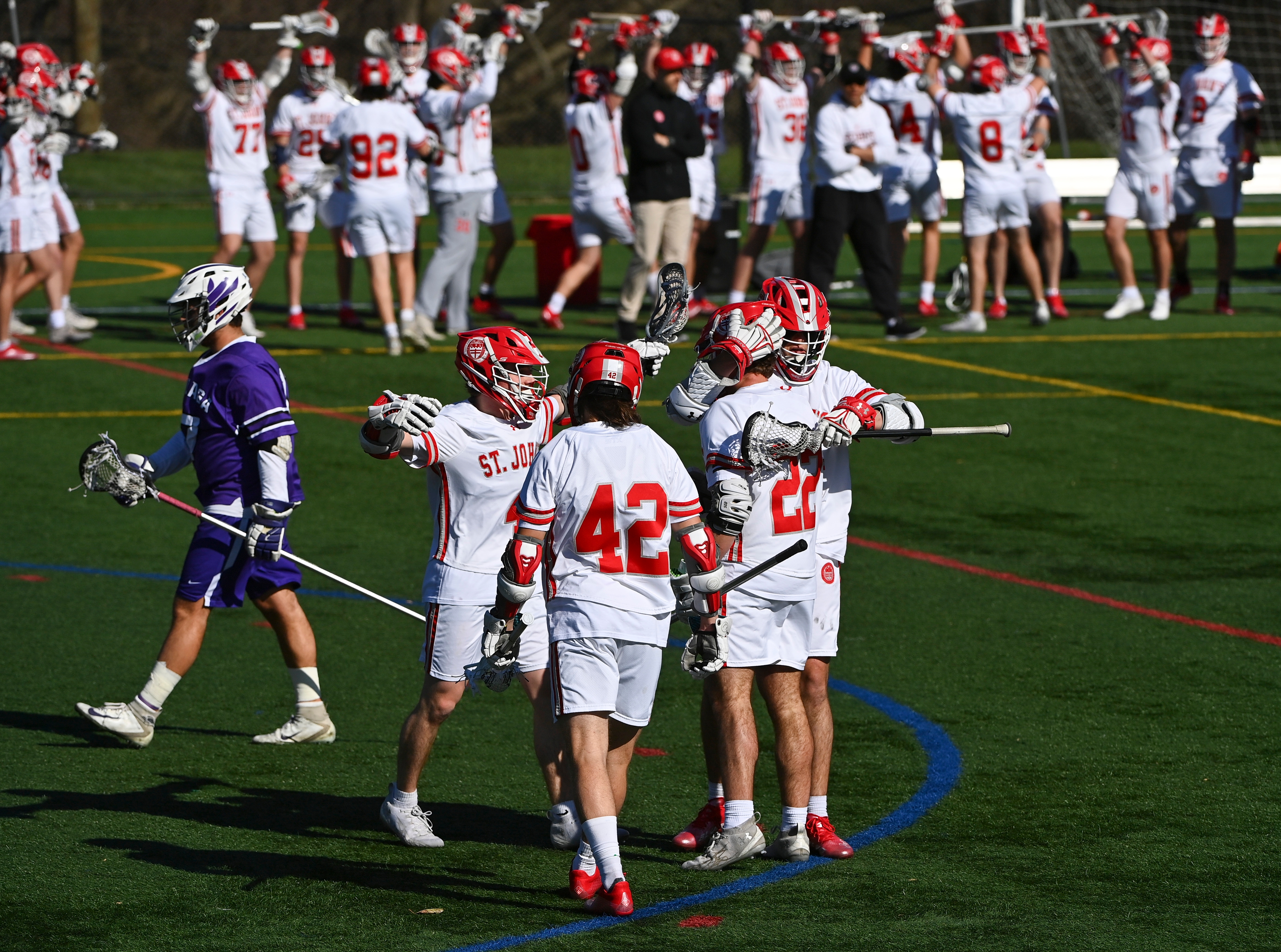 Lacrosse players in white jerseys celebrate on the field while a player in a purple jersey walks by. Other team members observe in the background