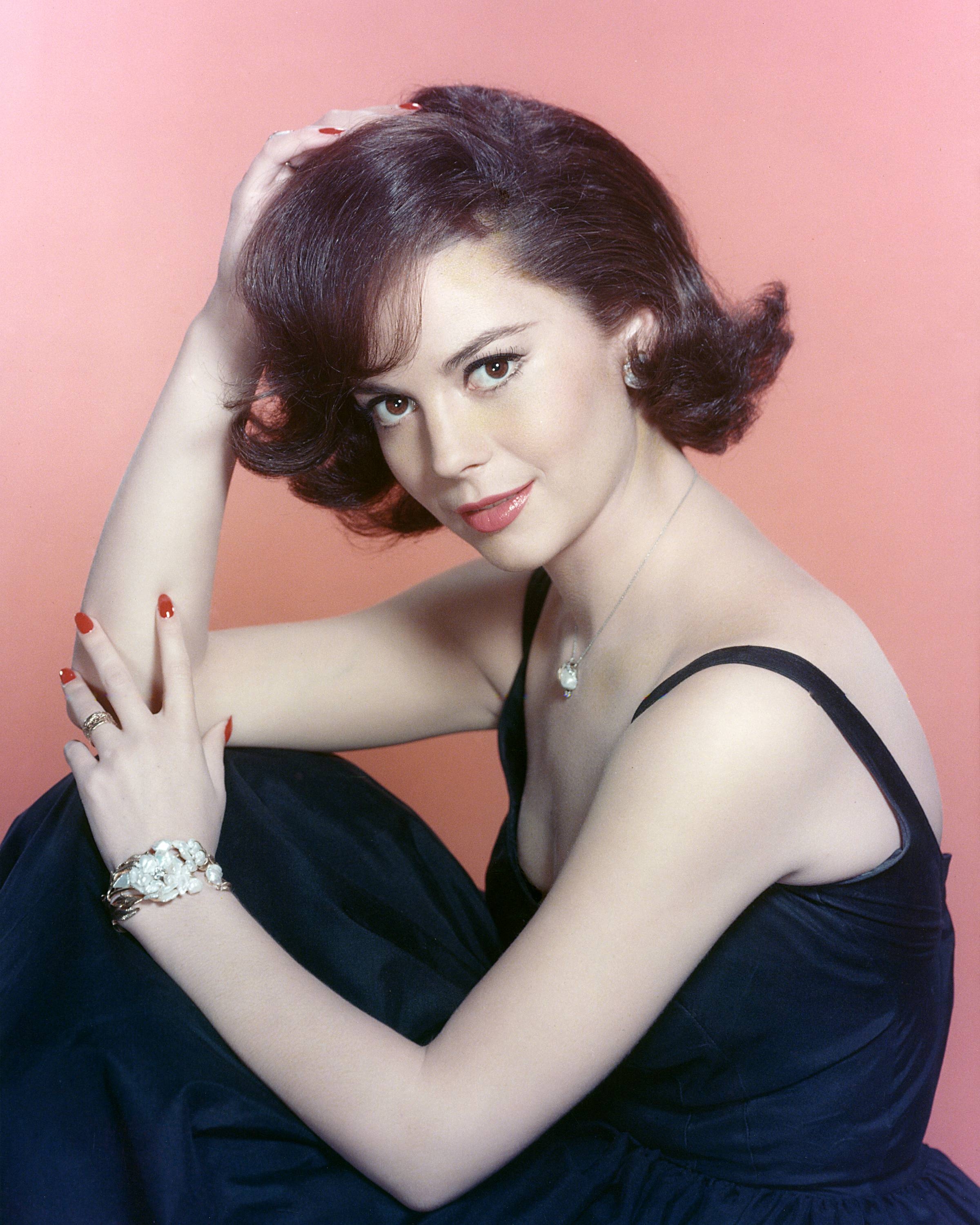Person posing elegantly, wearing a classic black dress and jewelry against a plain backdrop, hand resting softly on head