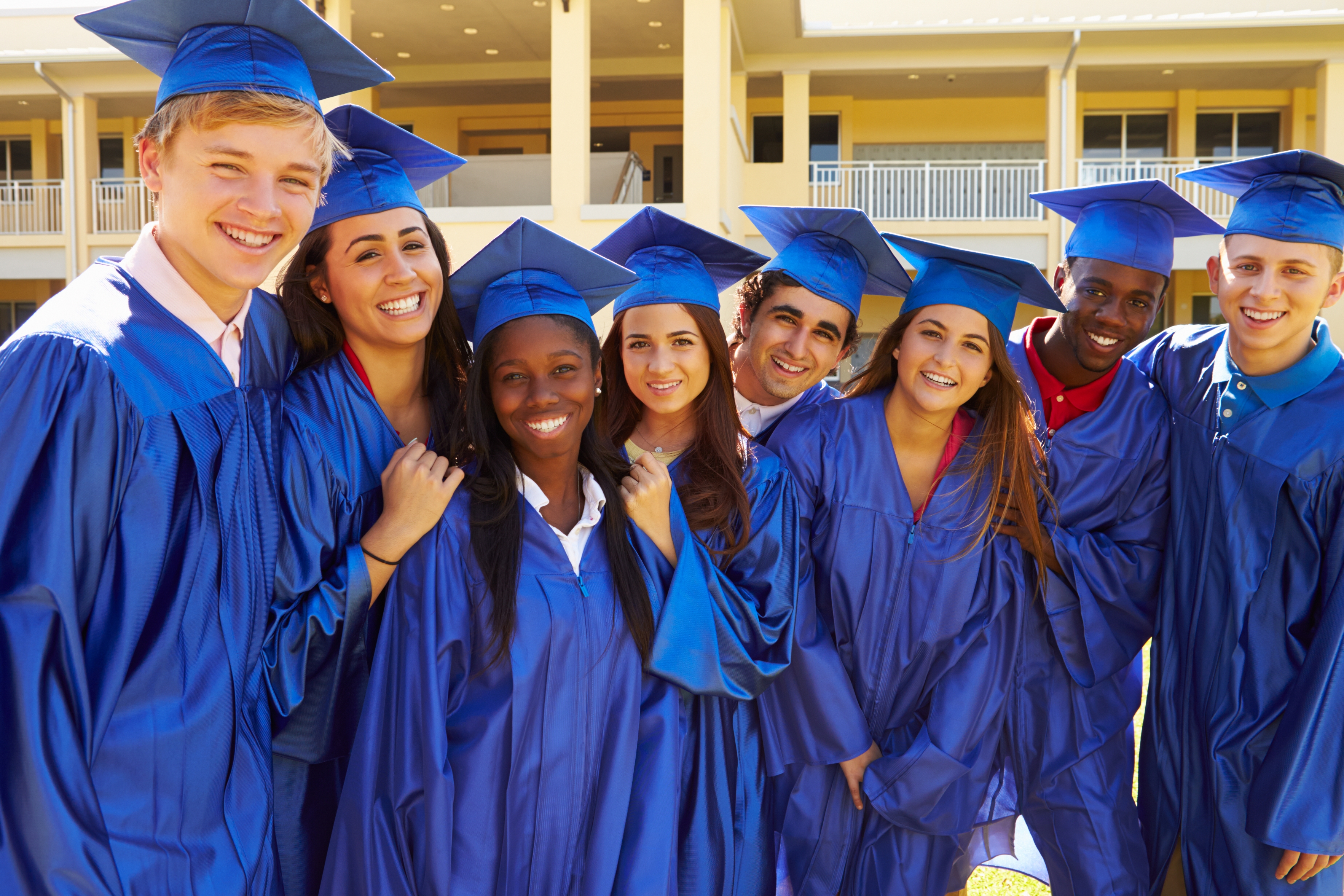 Group of graduates in blue caps and gowns, smiling and posing closely together outside a building