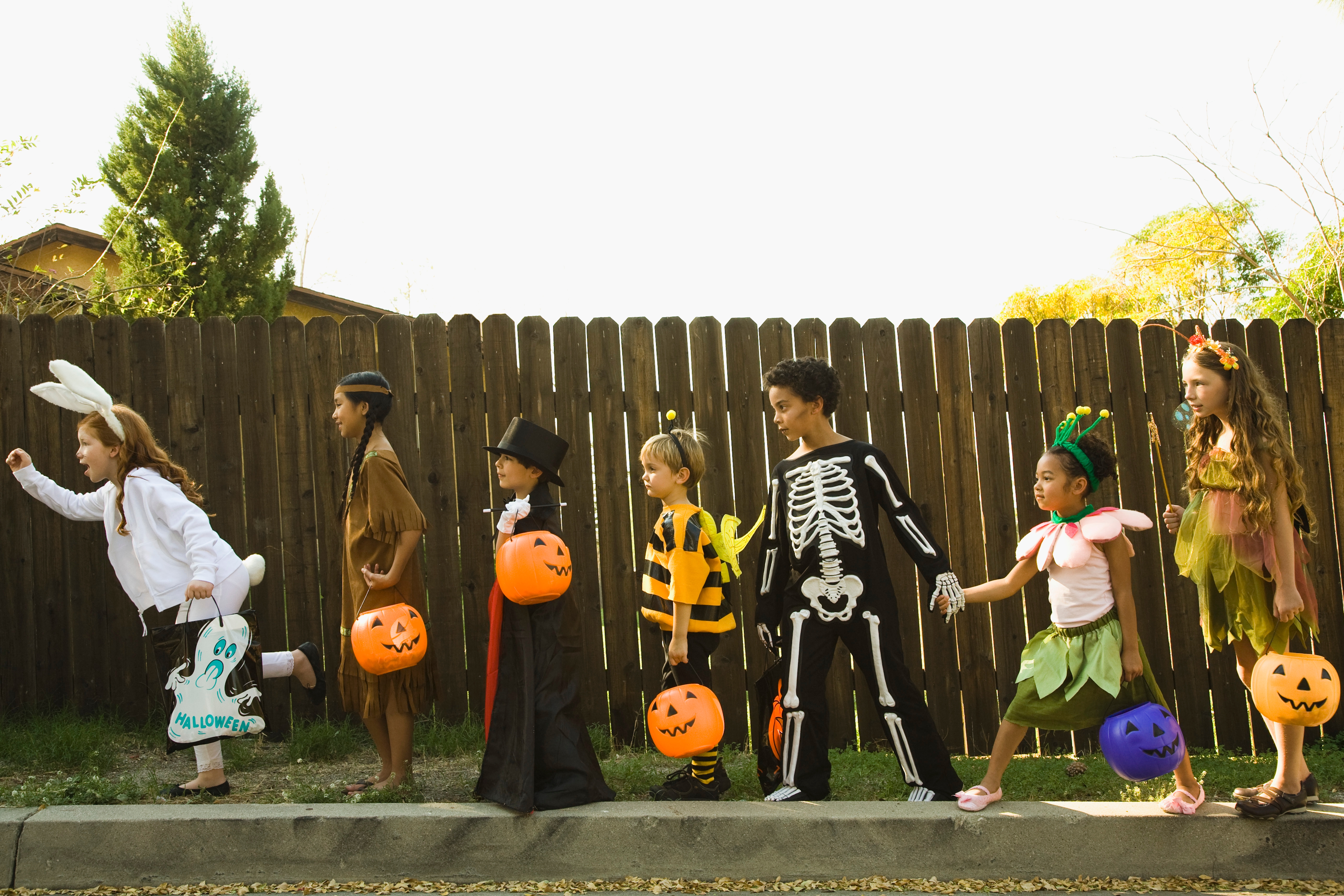 Children in Halloween costumes, including a bunny, cowboy, witch, bee, skeleton, fairy, and pirate, walk in line holding pumpkin baskets
