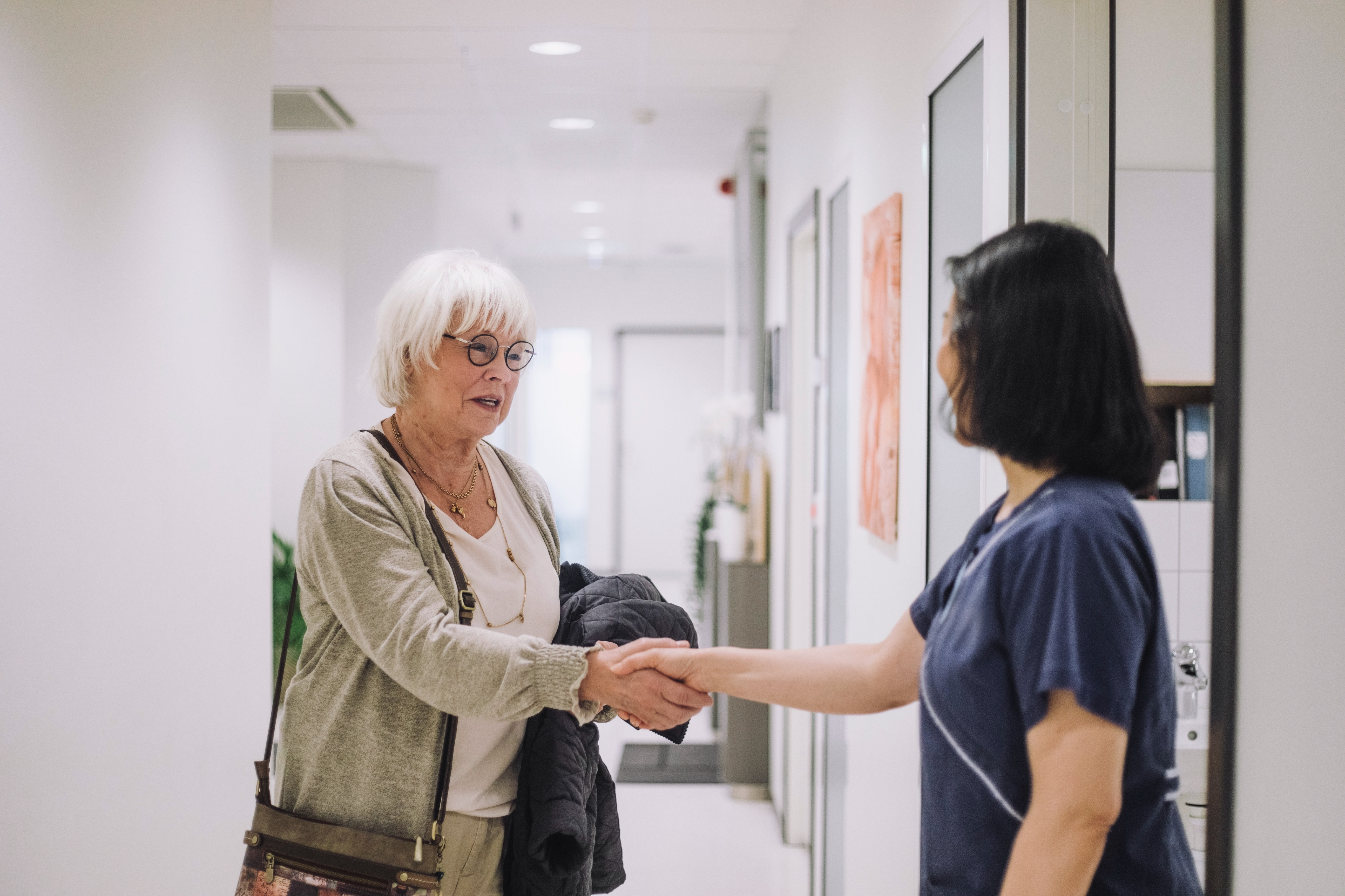 An older woman with a shoulder bag shakes hands with a healthcare worker in a hallway