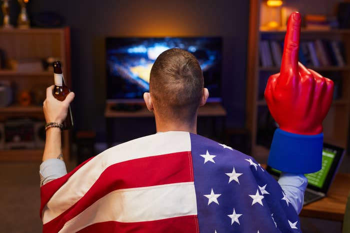 Person with an American flag draped over their shoulders, holding a foam finger and beer, watching a sports game on a TV in a cozy room