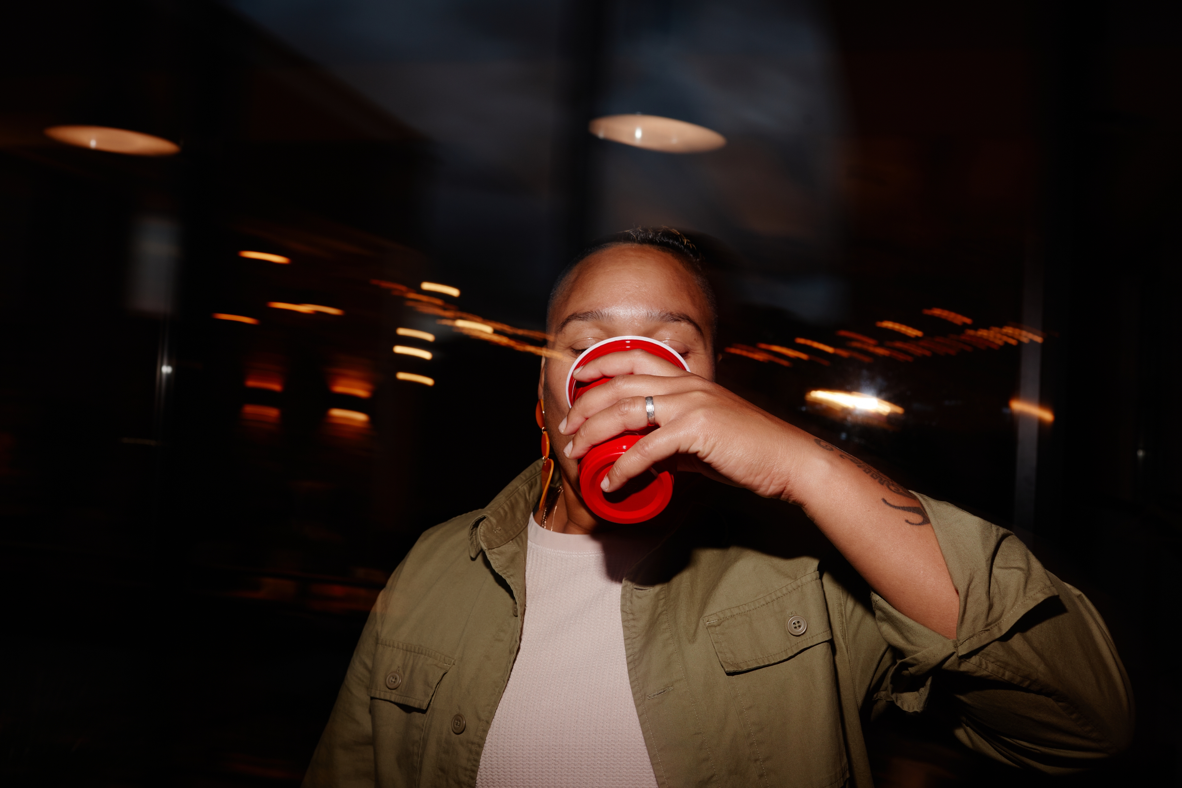 Person in a casual jacket holds a red cup to their face, drinking. Blurred background suggests movement or a party setting