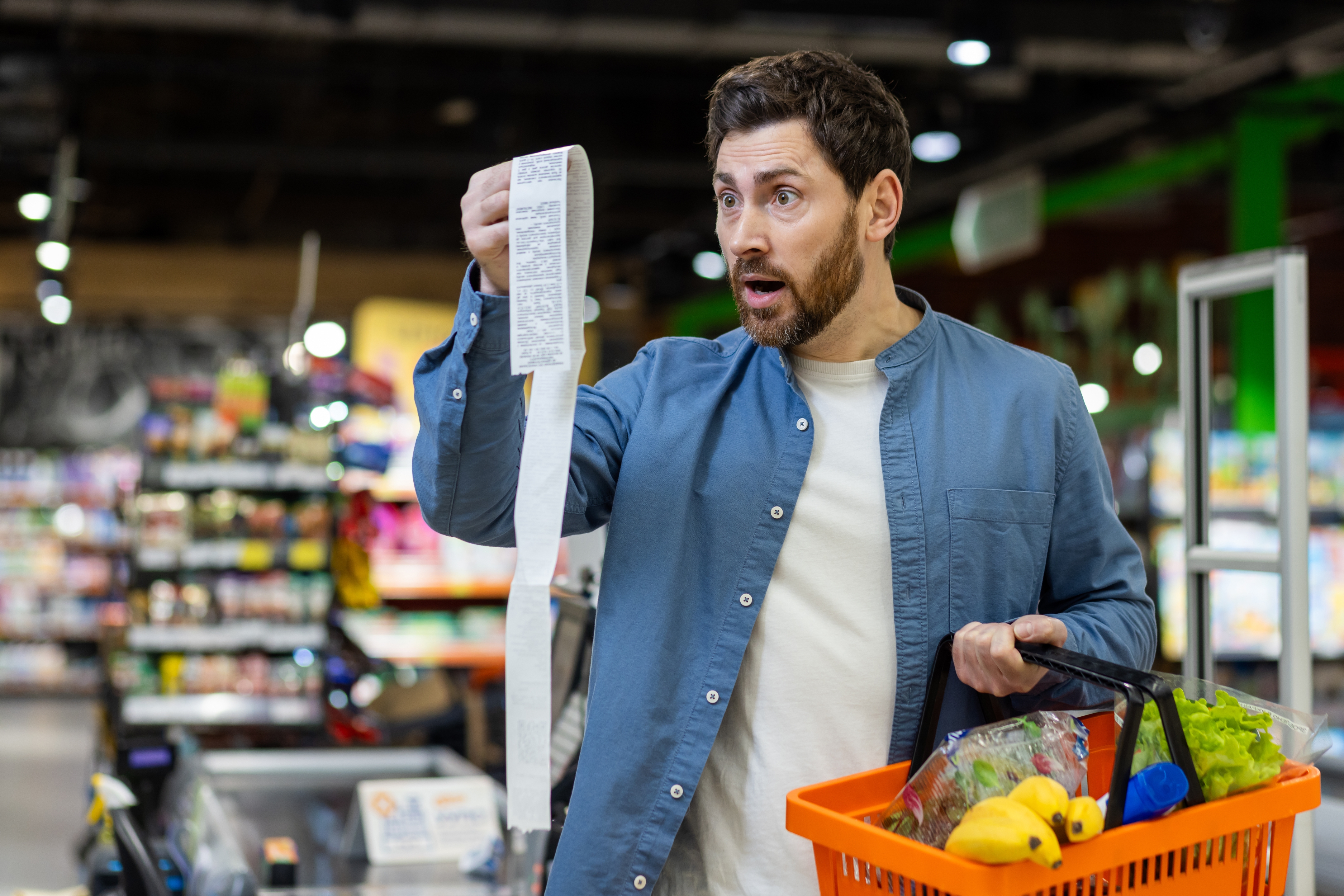 Man in grocery store looks surprised at a long receipt while holding a basket of groceries