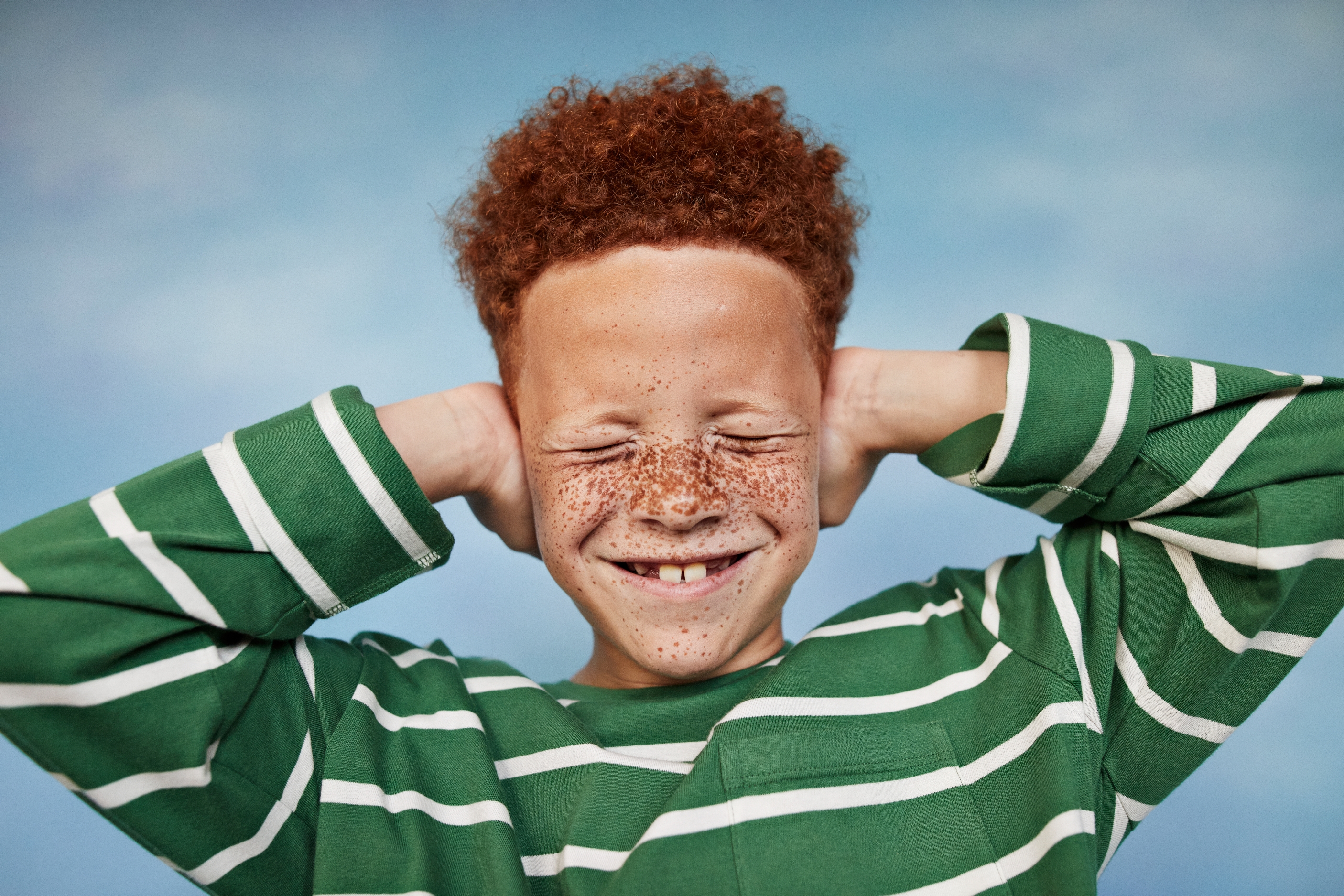 Child with curly hair and freckles smiles with eyes closed, wearing a green striped shirt, covering ears playfully in an outdoor setting