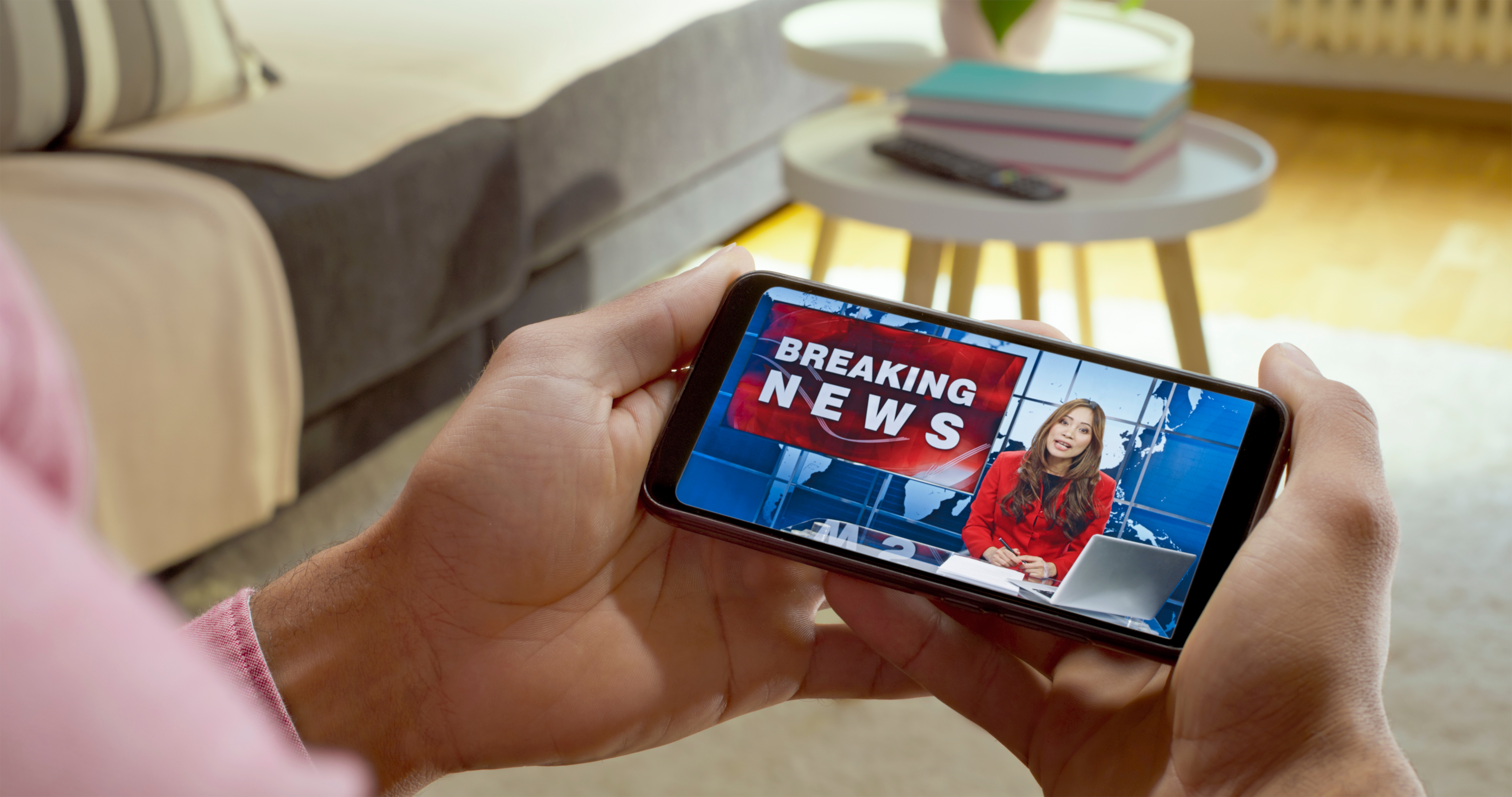 Person watches breaking news on a smartphone, with a news anchor at a desk onscreen. The background shows a bright living room
