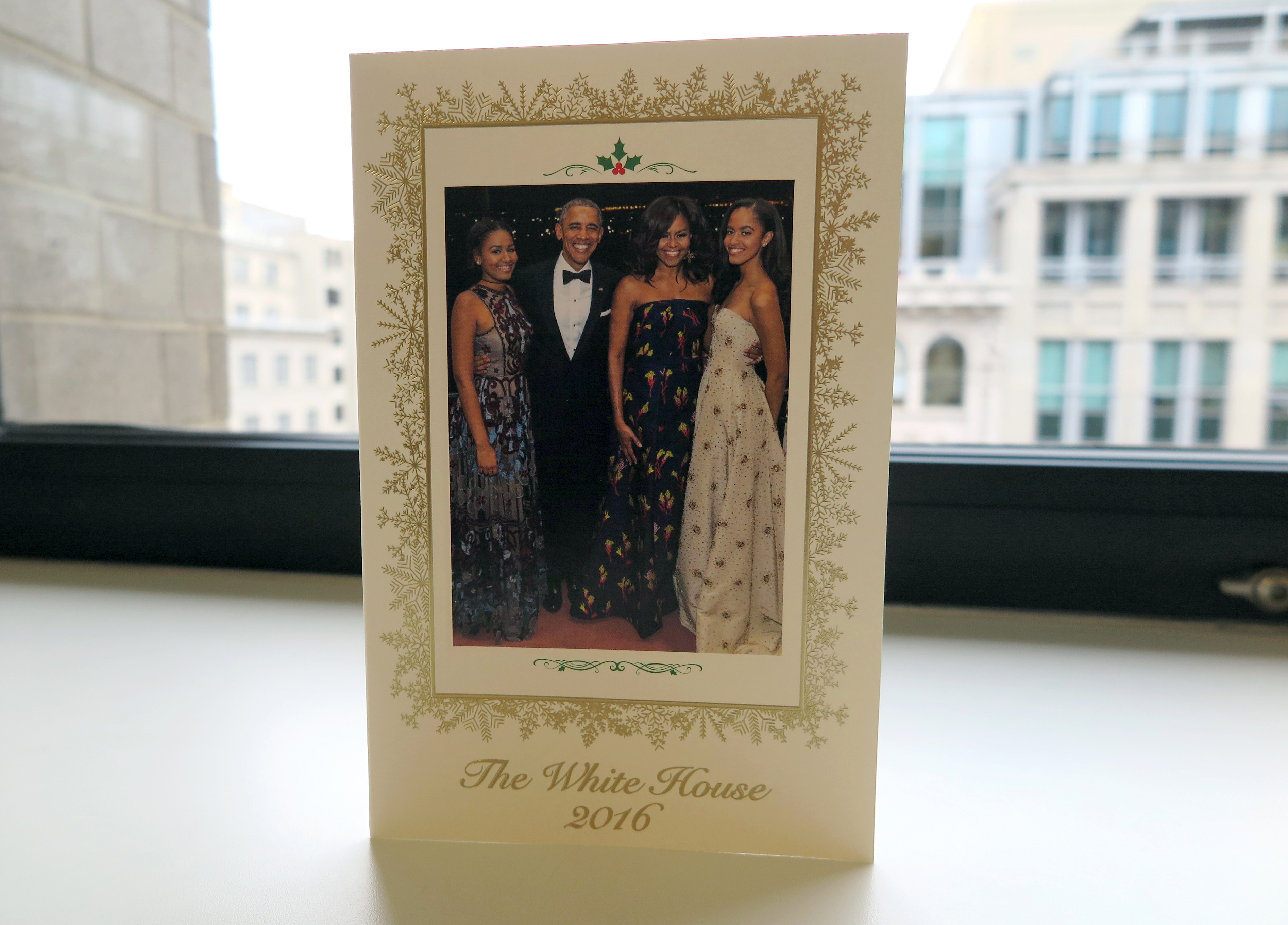 Family photo on a greeting card with "The White House 2016" printed below, showing formal attire. Image placed on a window sill with buildings outside