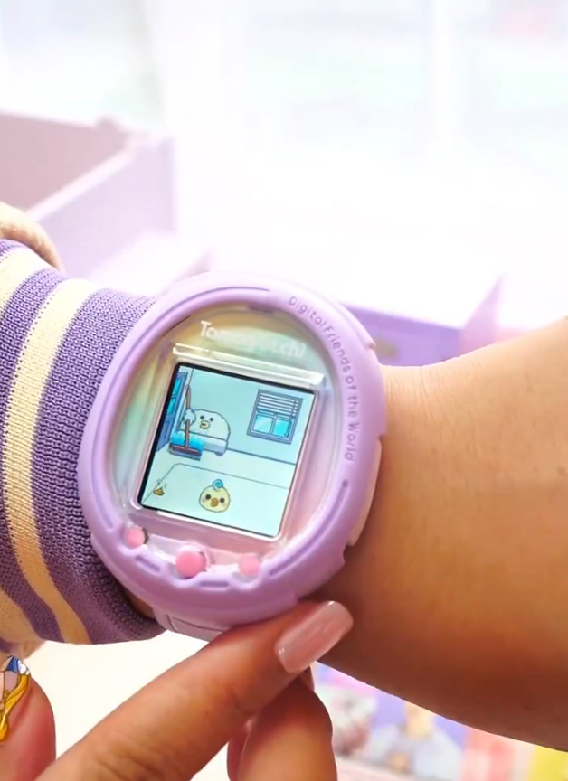 A person wears a digital pet watch, displaying a virtual room and character. Caption below reads, “It was like $125.”