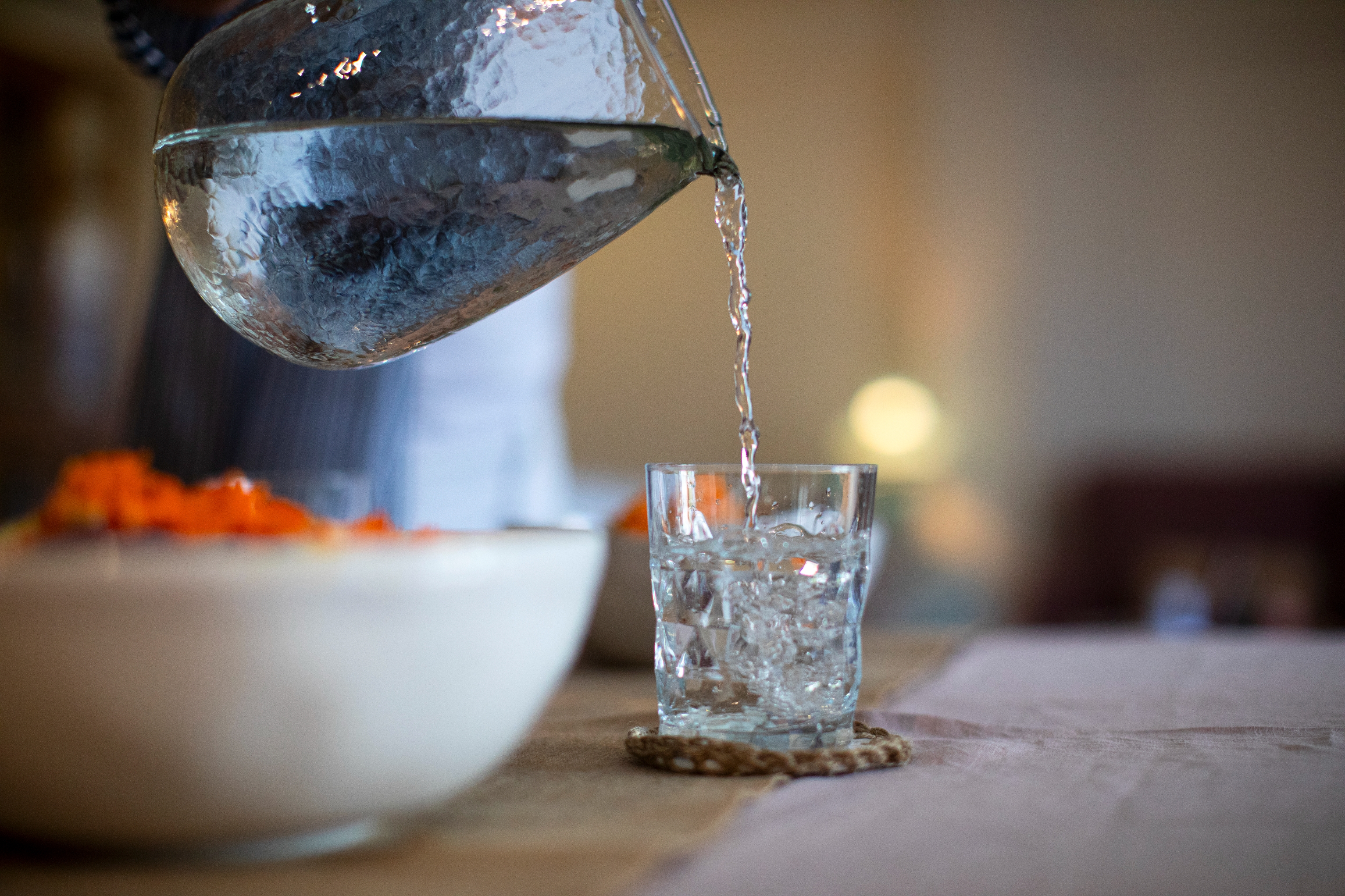 Water from a clear pitcher is being poured into a glass on a table next to a bowl