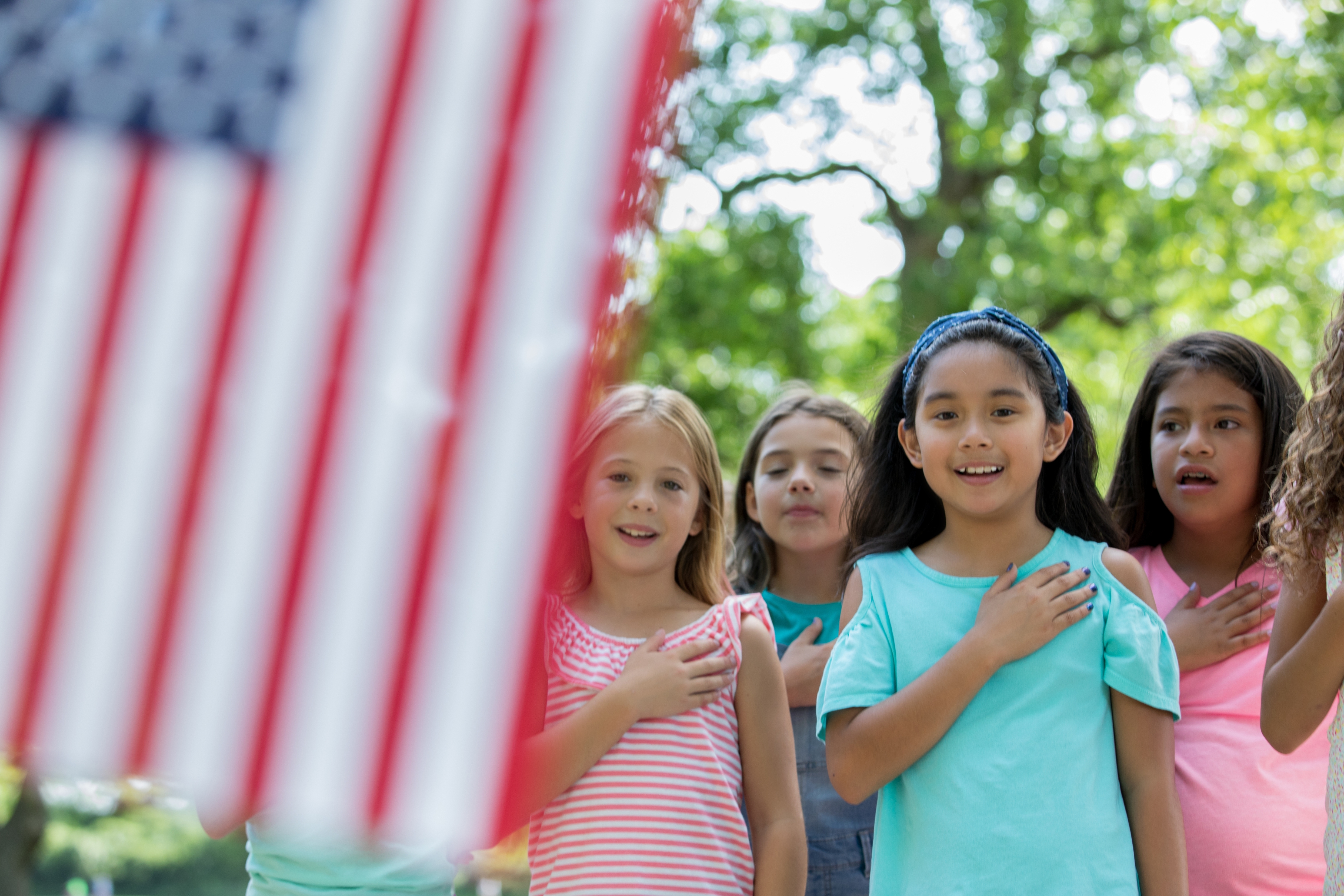 Children stand outdoors with hands on hearts, facing a blurred American flag, appearing to recite the Pledge of Allegiance