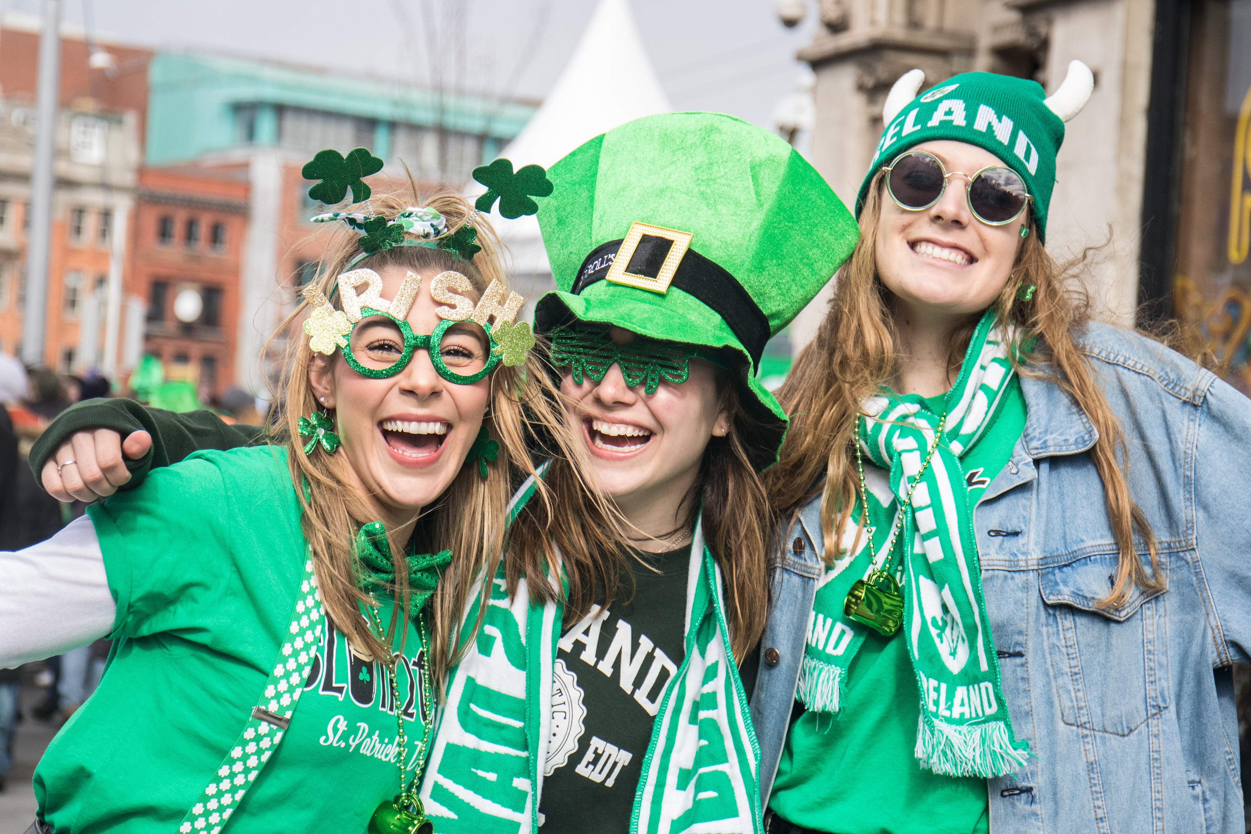 Three people celebrate St. Patrick's Day wearing themed hats, glasses, and green attire, smiling and outdoors