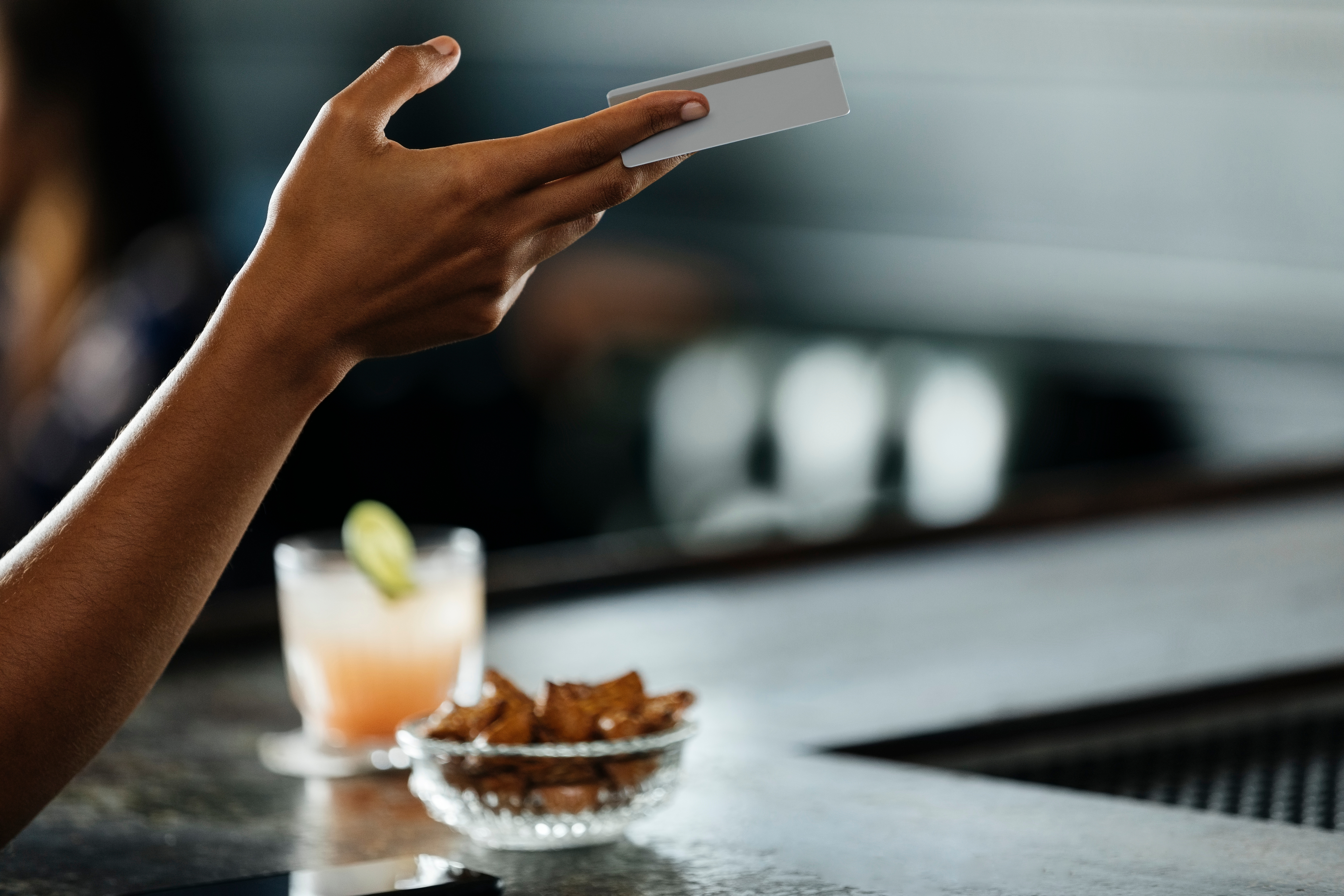 A person holds a smartphone at a bar with a cocktail and a bowl of snacks on the counter
